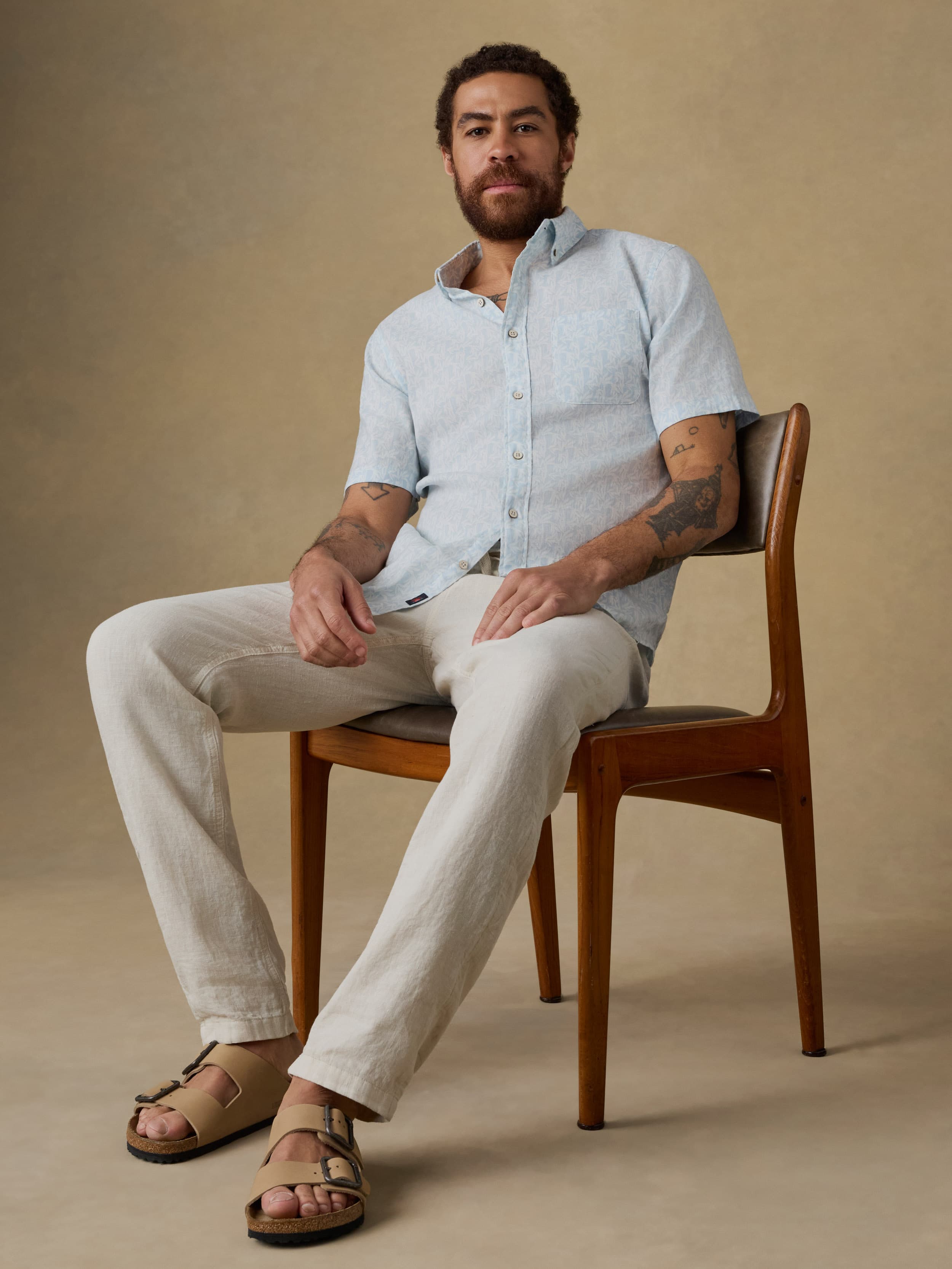 Man sitting on a wooden chair wearing a light blue linen shirt and beige linen pants against a beige background