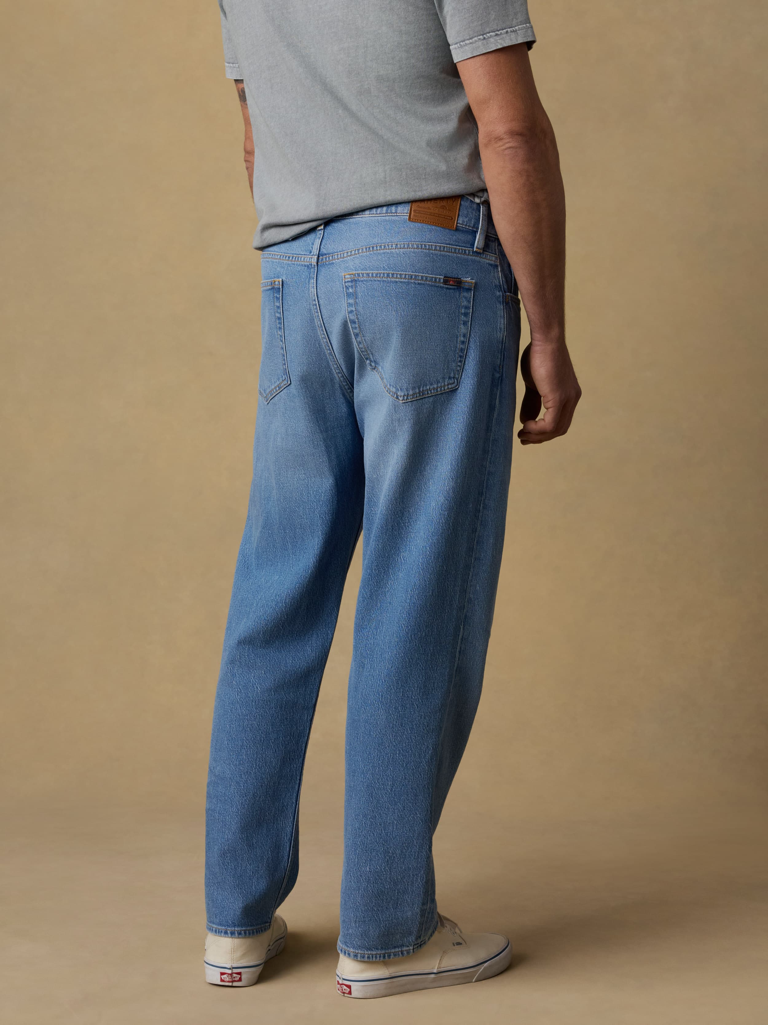 Back view of a man wearing a gray short sleeve t-shirt and blue jeans against a beige background