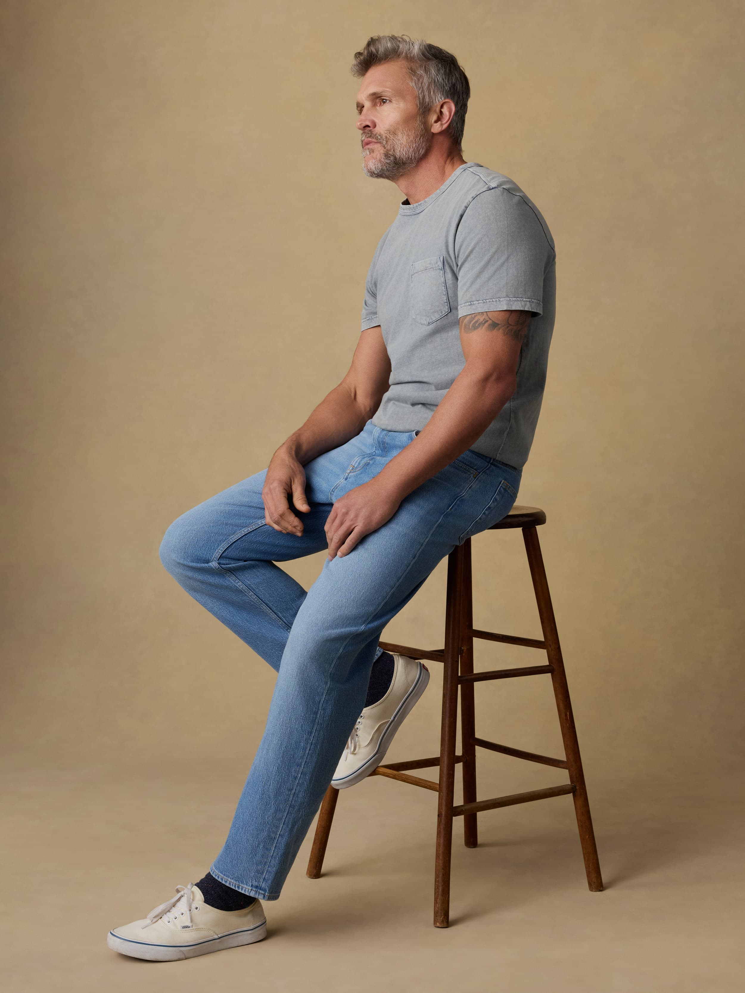 Man sitting on a wooden stool wearing a gray short sleeve t-shirt and blue jeans against a beige background