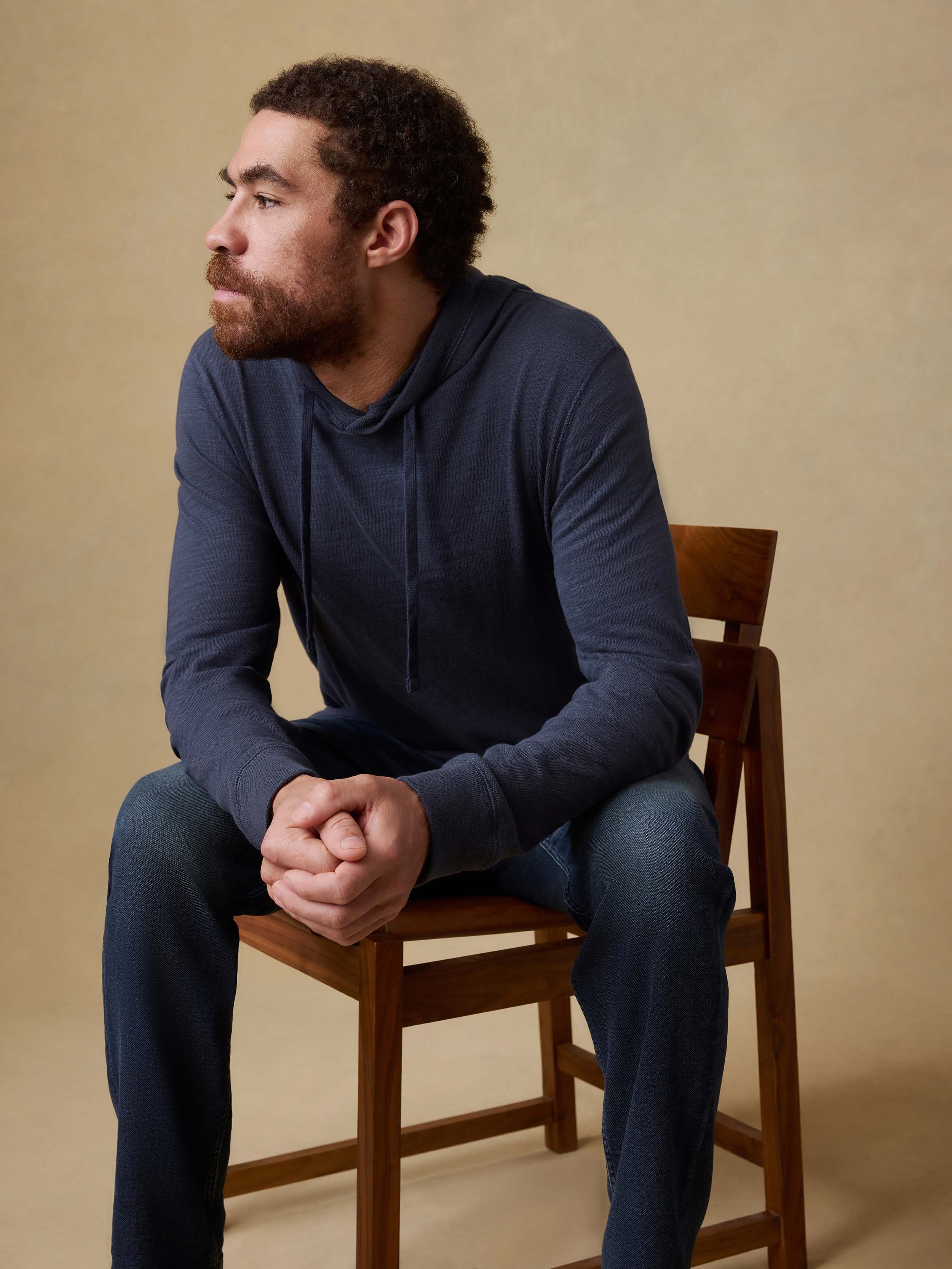 Man sitting on a wooden chair wearing a blue hoodie and blue jeans against a beige background