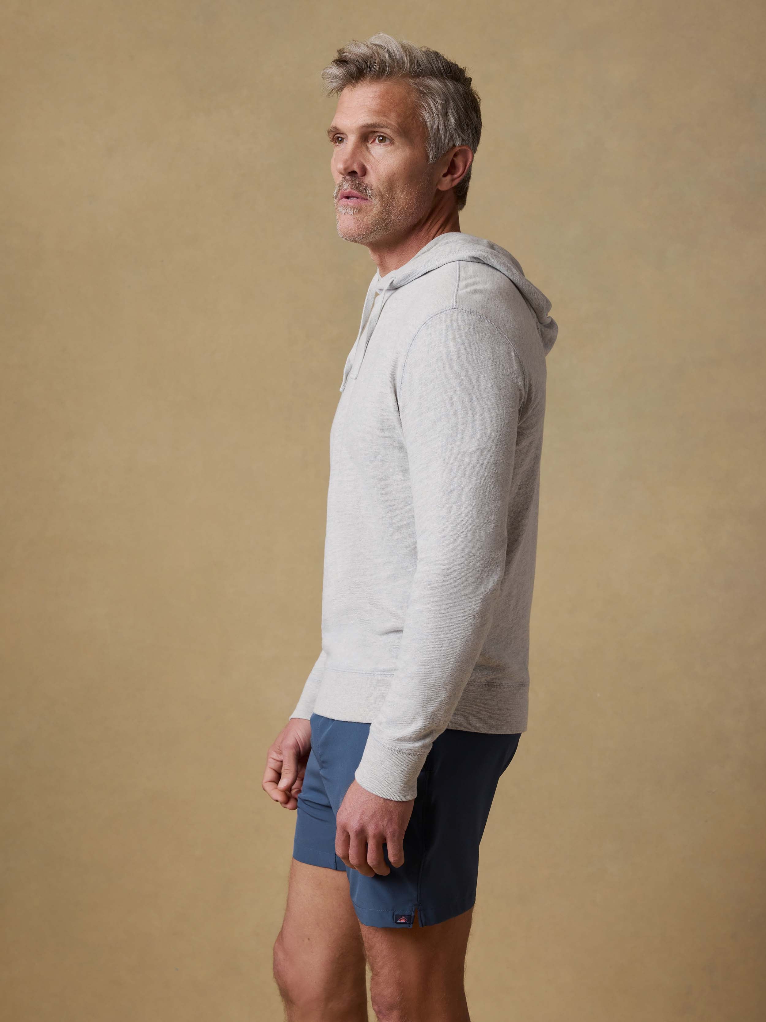 Man wearing a light gray hoodie and blue shorts against a beige background