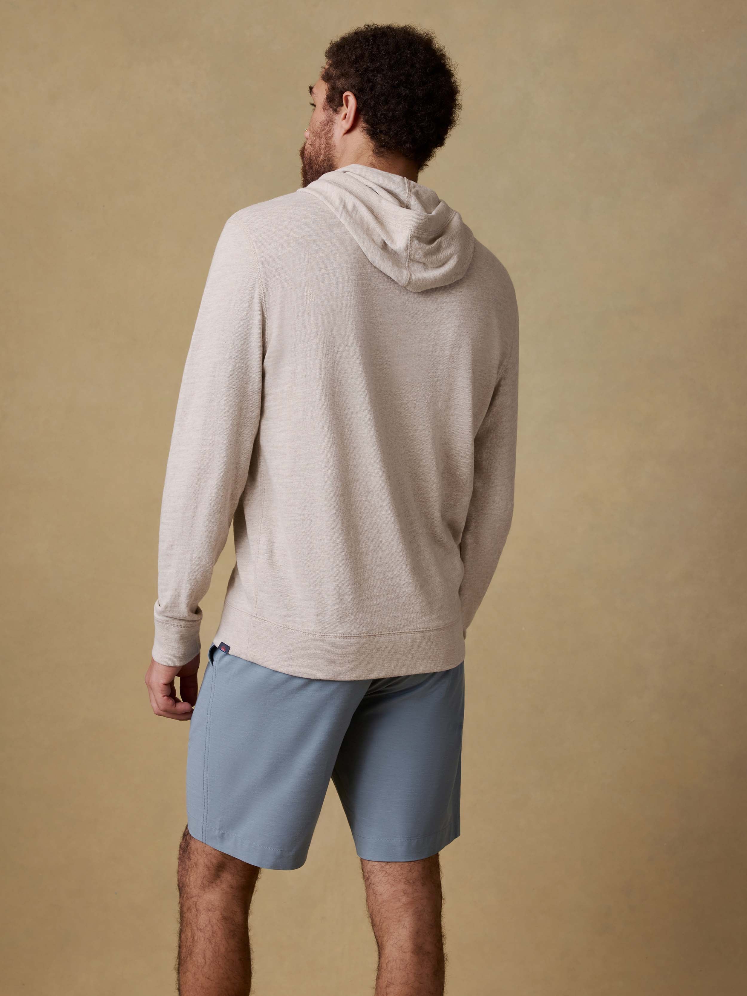 Back view of a man wearing a beige hoodie and light blue shorts against a beige background