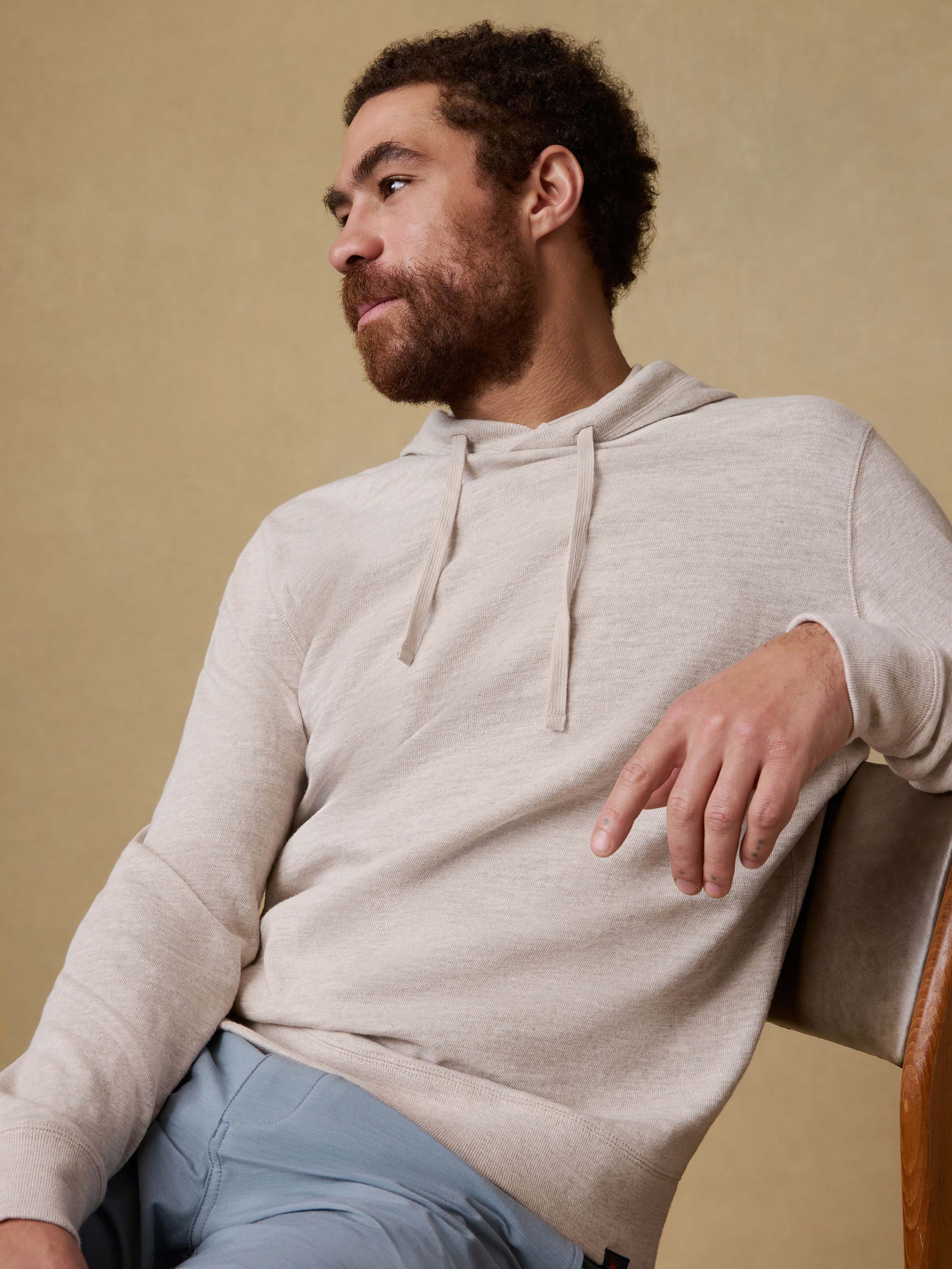 Man wearing a beige hoodie and blue shorts sitting on a chair against a beige background