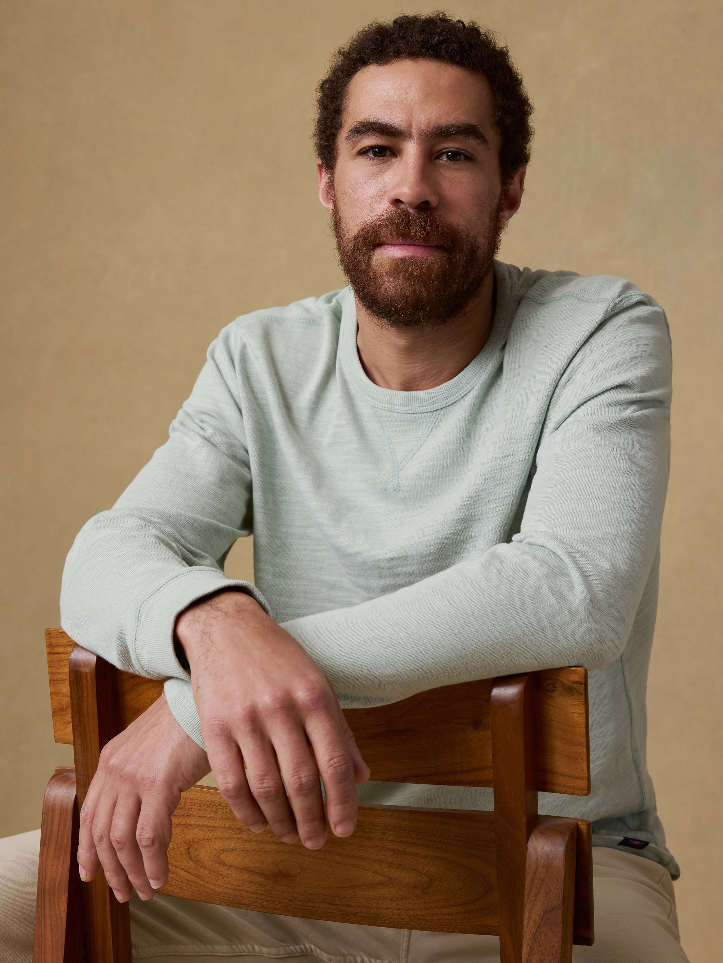 Man sitting on a wooden chair wearing a light green long sleeve shirt against a beige background