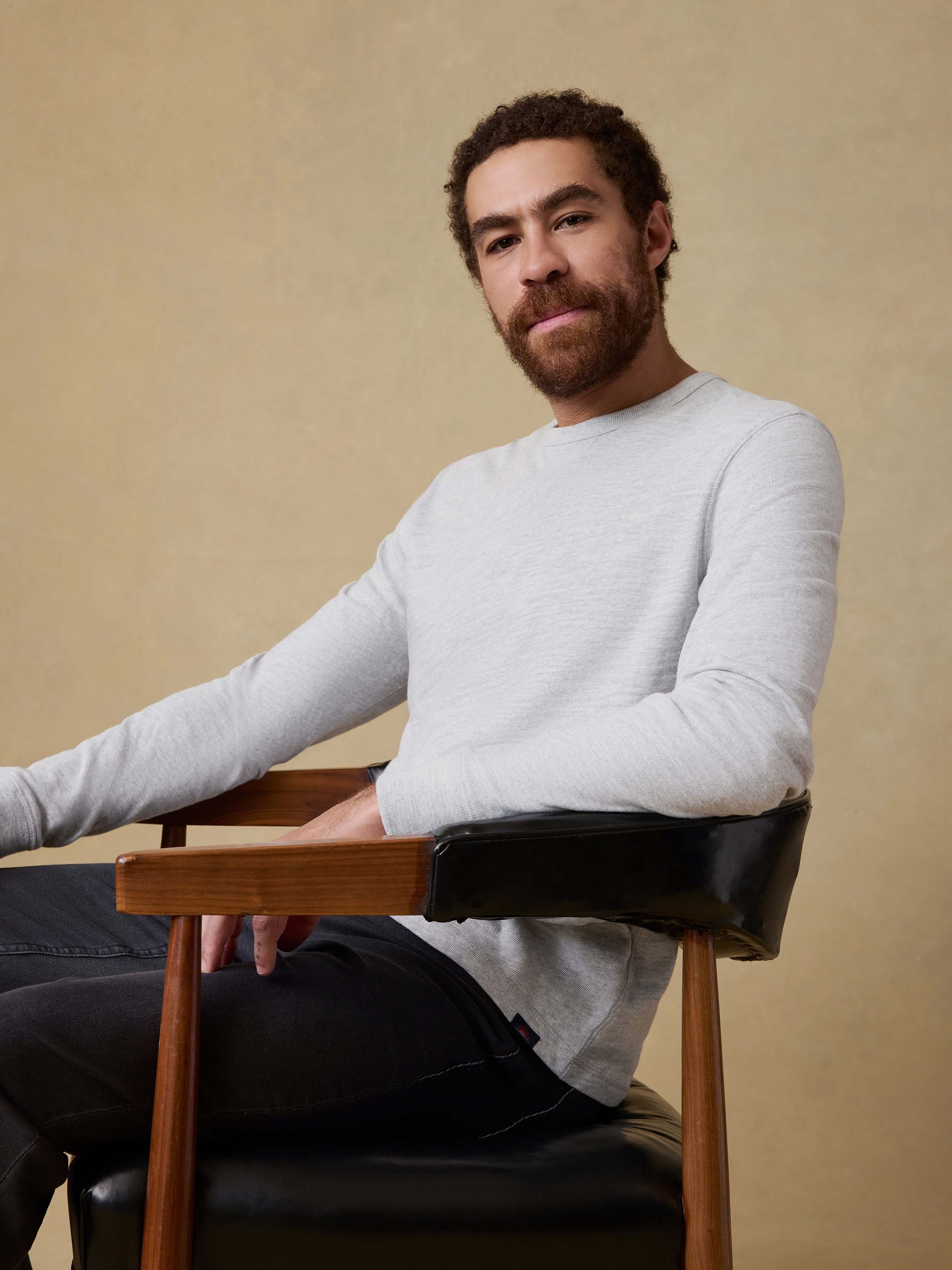 Man sitting in a chair wearing a light gray long sleeve shirt and dark pants against a beige background