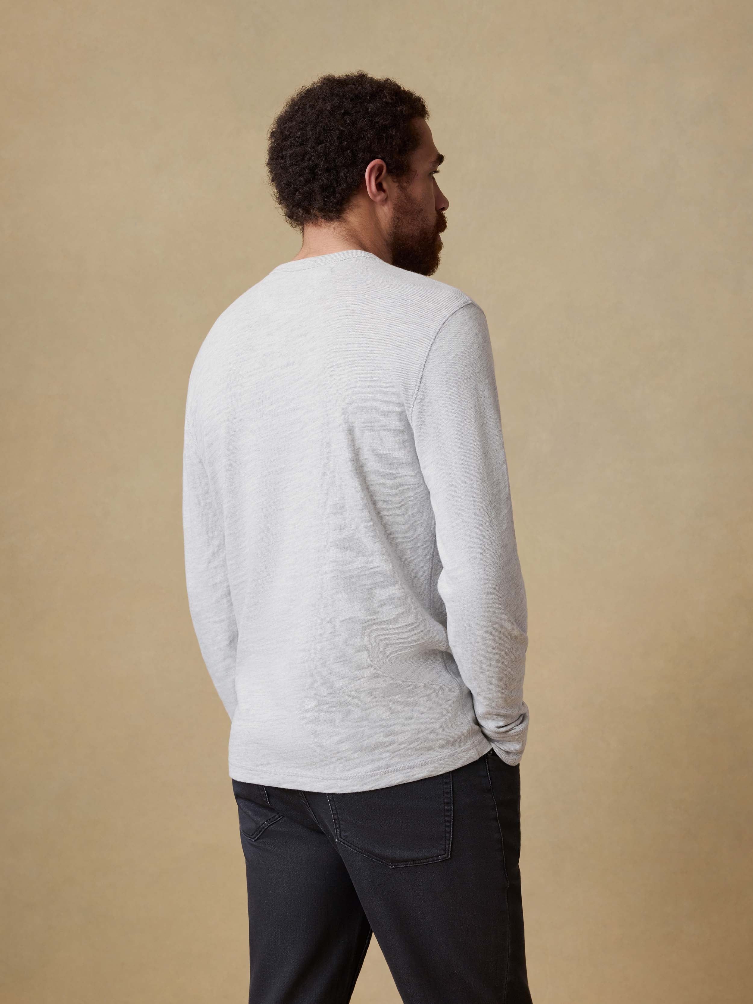 Back view of a man wearing a light gray long sleeve shirt and dark pants against a beige background