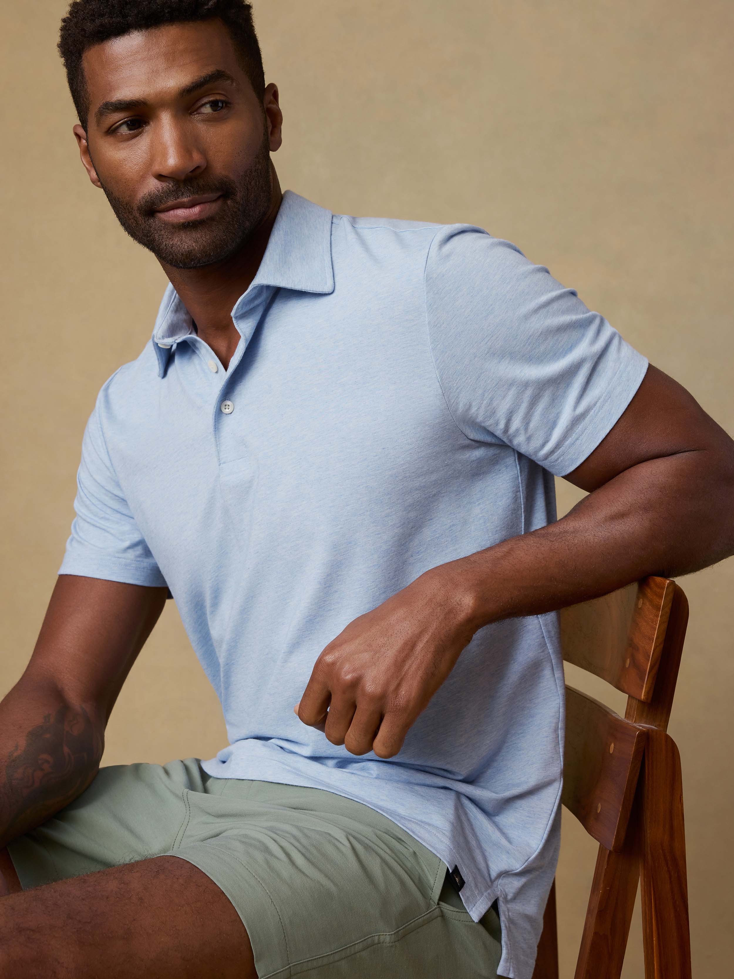 Man wearing a light blue polo shirt and green shorts sitting on a wooden chair against a beige background