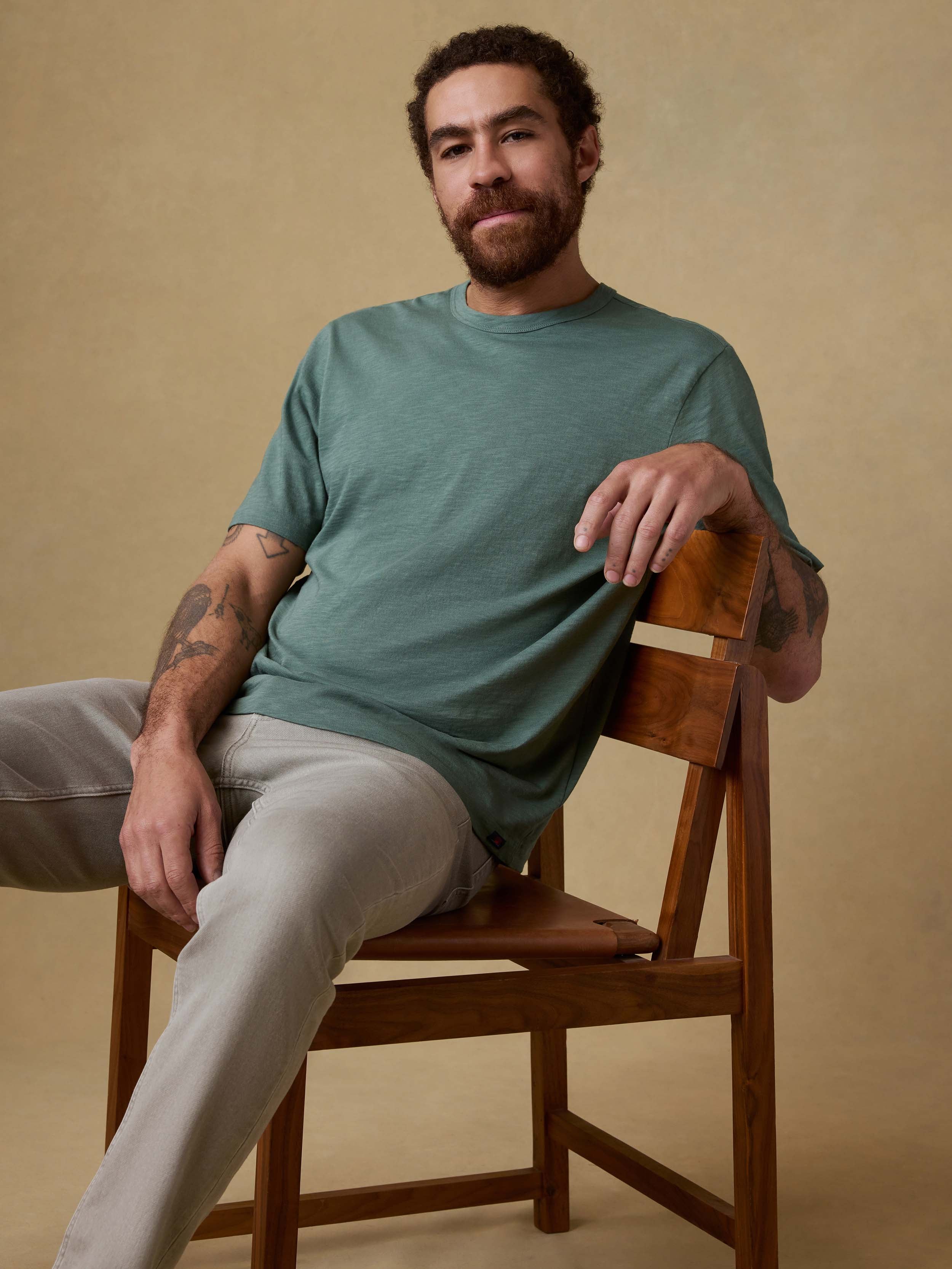 Man sitting on a wooden chair wearing a green t-shirt and light gray pants against a beige background