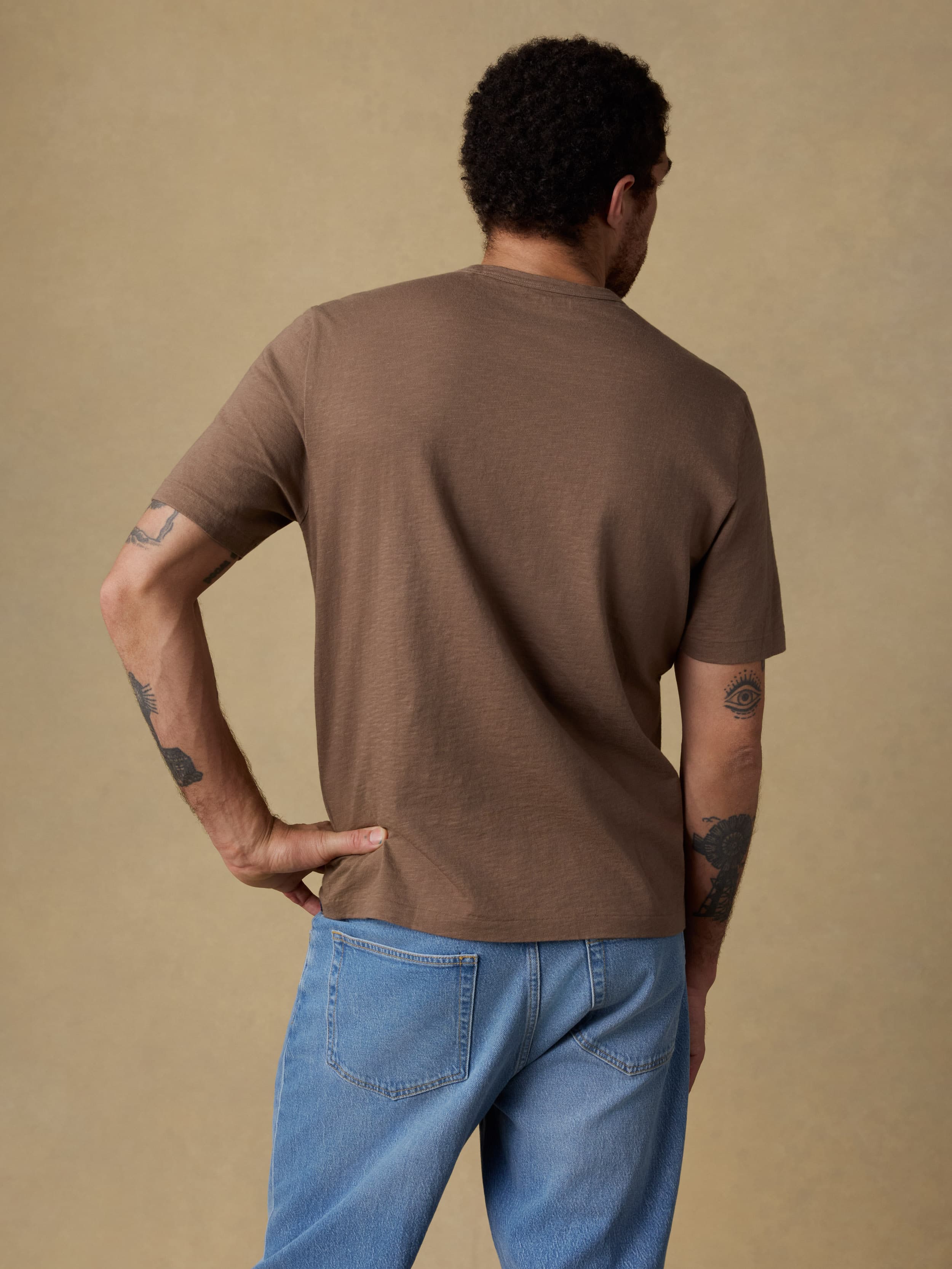 Back view of a man wearing a brown short sleeve t-shirt and blue jeans against a beige background