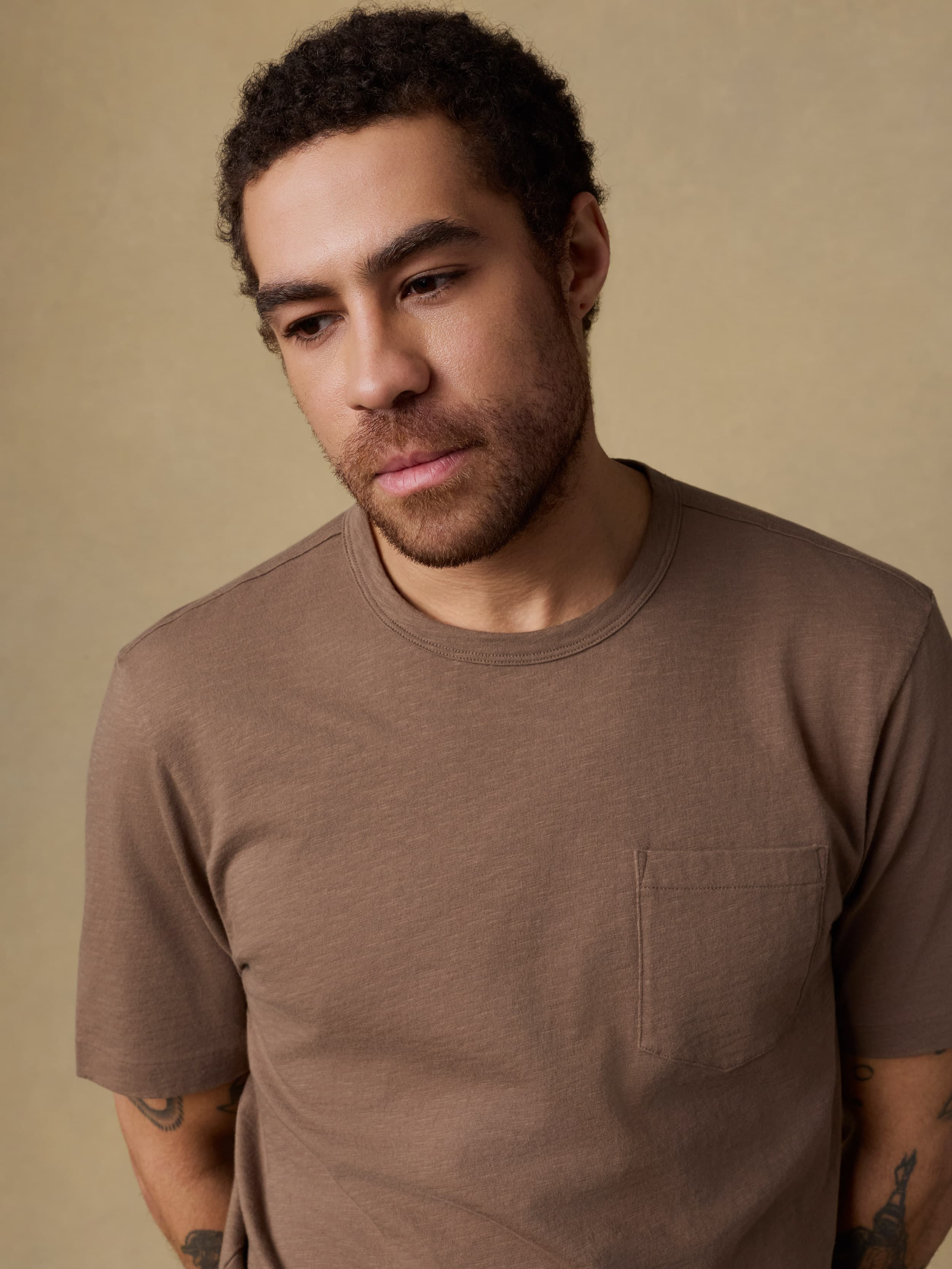 Man wearing a brown short sleeve t-shirt with a pocket on the left chest against a beige background
