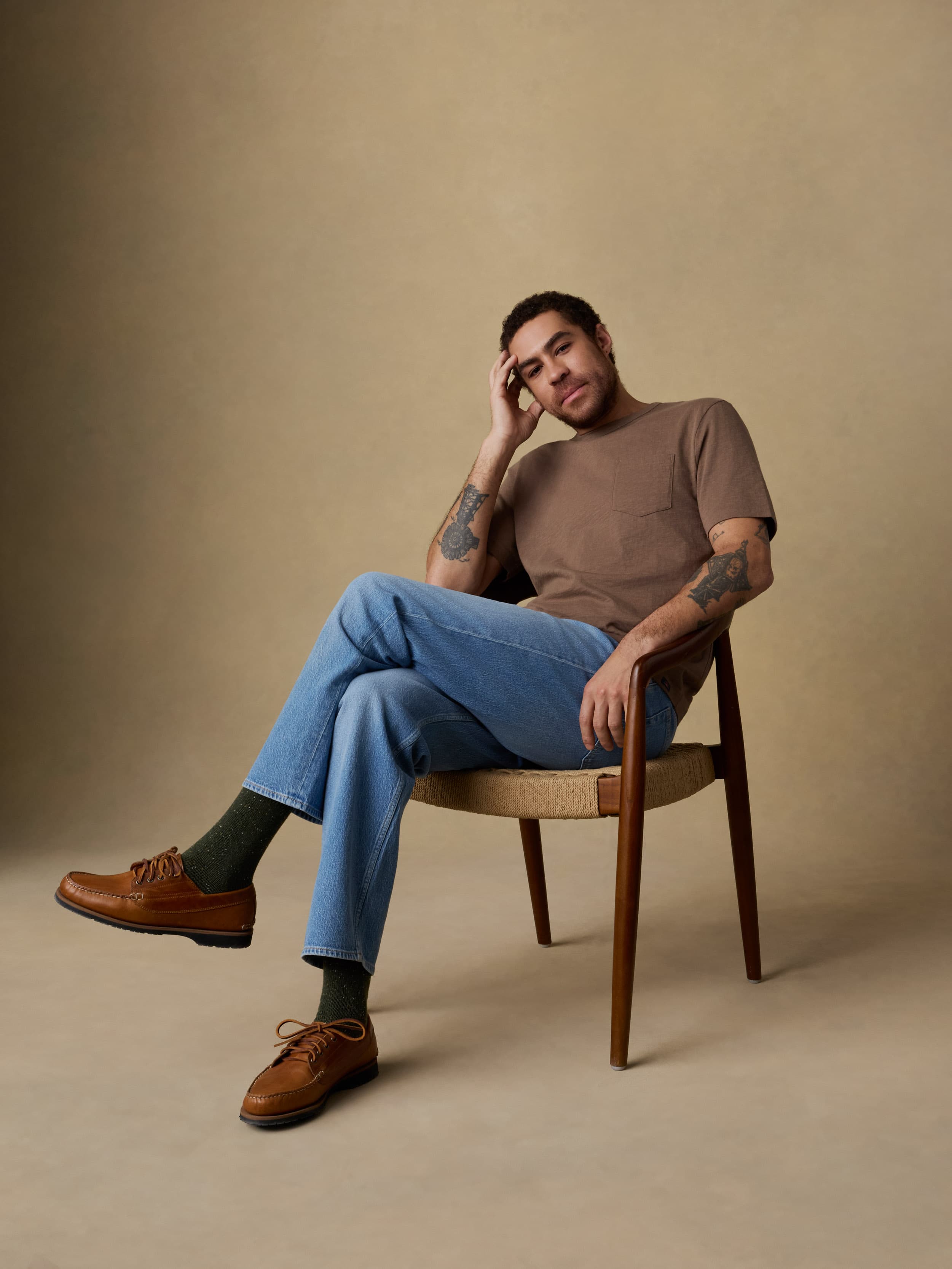 Man sitting on a wooden chair wearing a brown short sleeve t-shirt, blue jeans, and brown shoes against a beige background