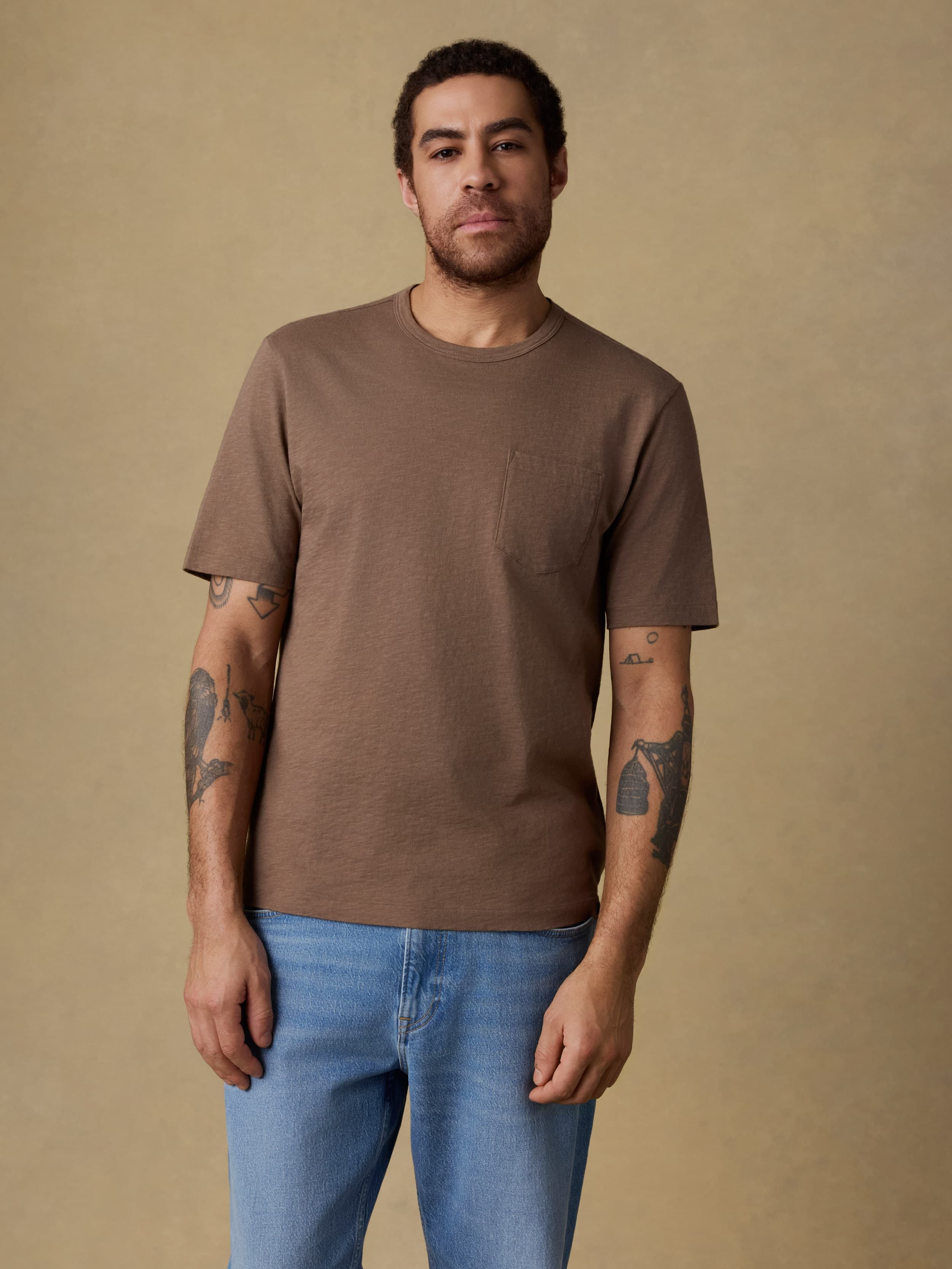 Man wearing a brown short sleeve t-shirt and blue jeans against a beige background