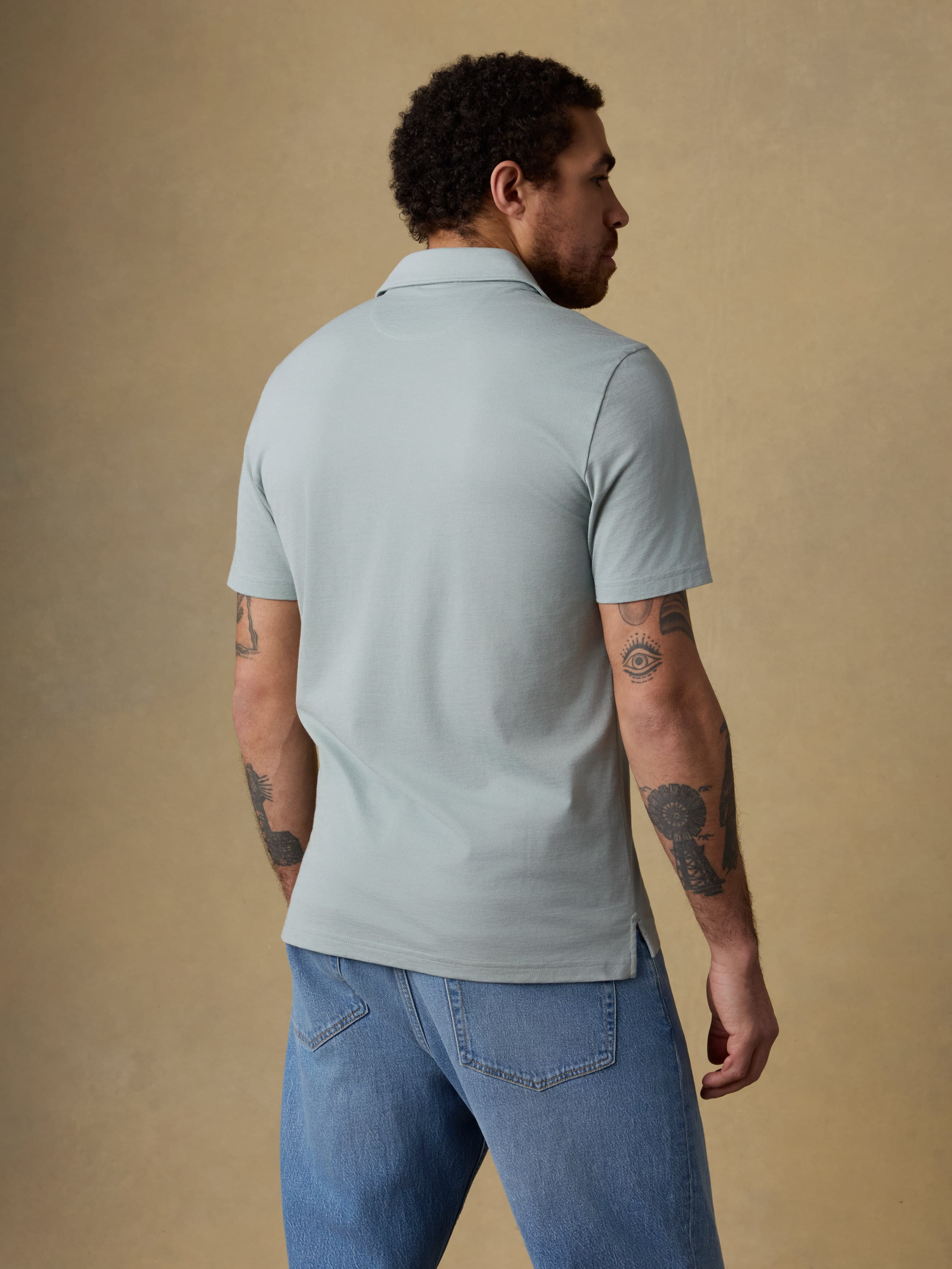 Back view of a man wearing a light green short sleeve polo shirt and blue jeans against a beige background