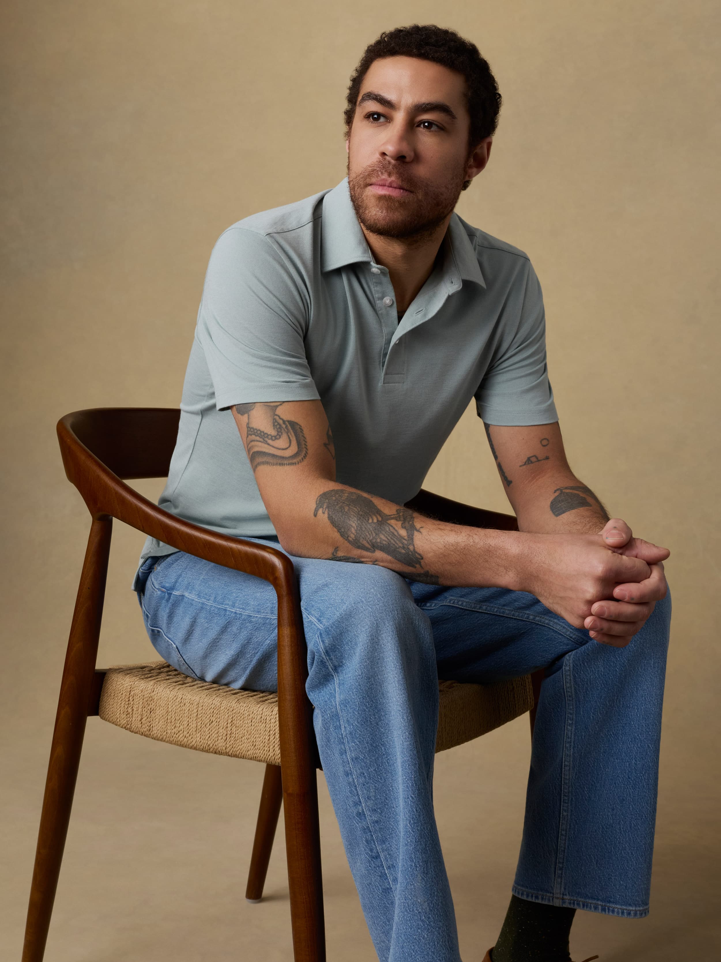 Man sitting on a wooden chair wearing a light green short sleeve polo shirt and blue jeans against a beige background