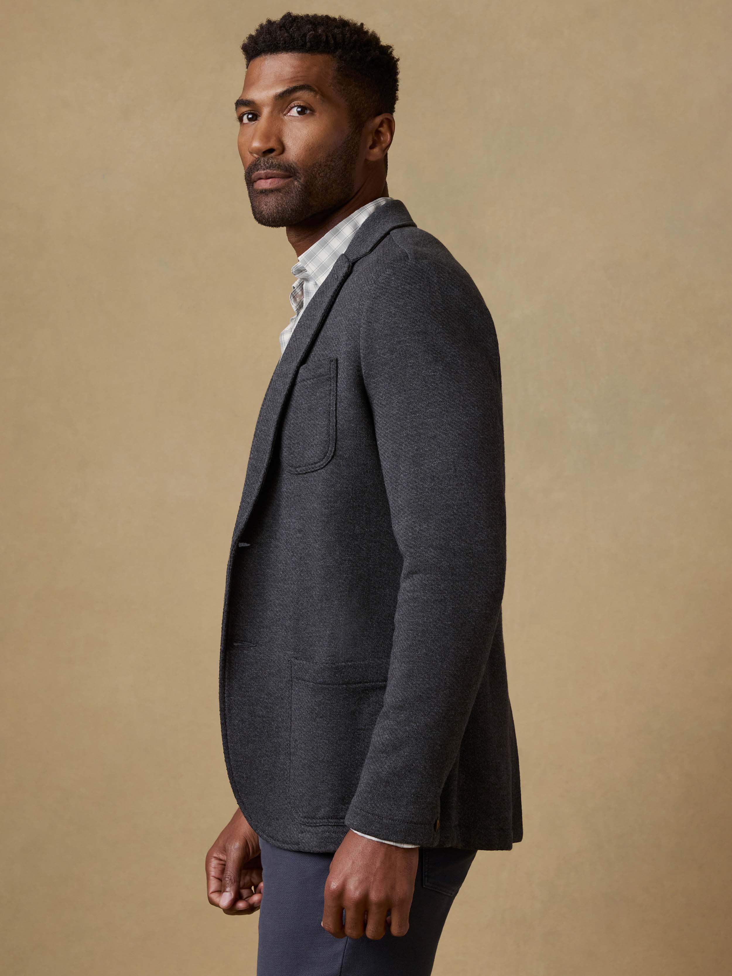 Man wearing a dark gray blazer over a plaid shirt with a beige background