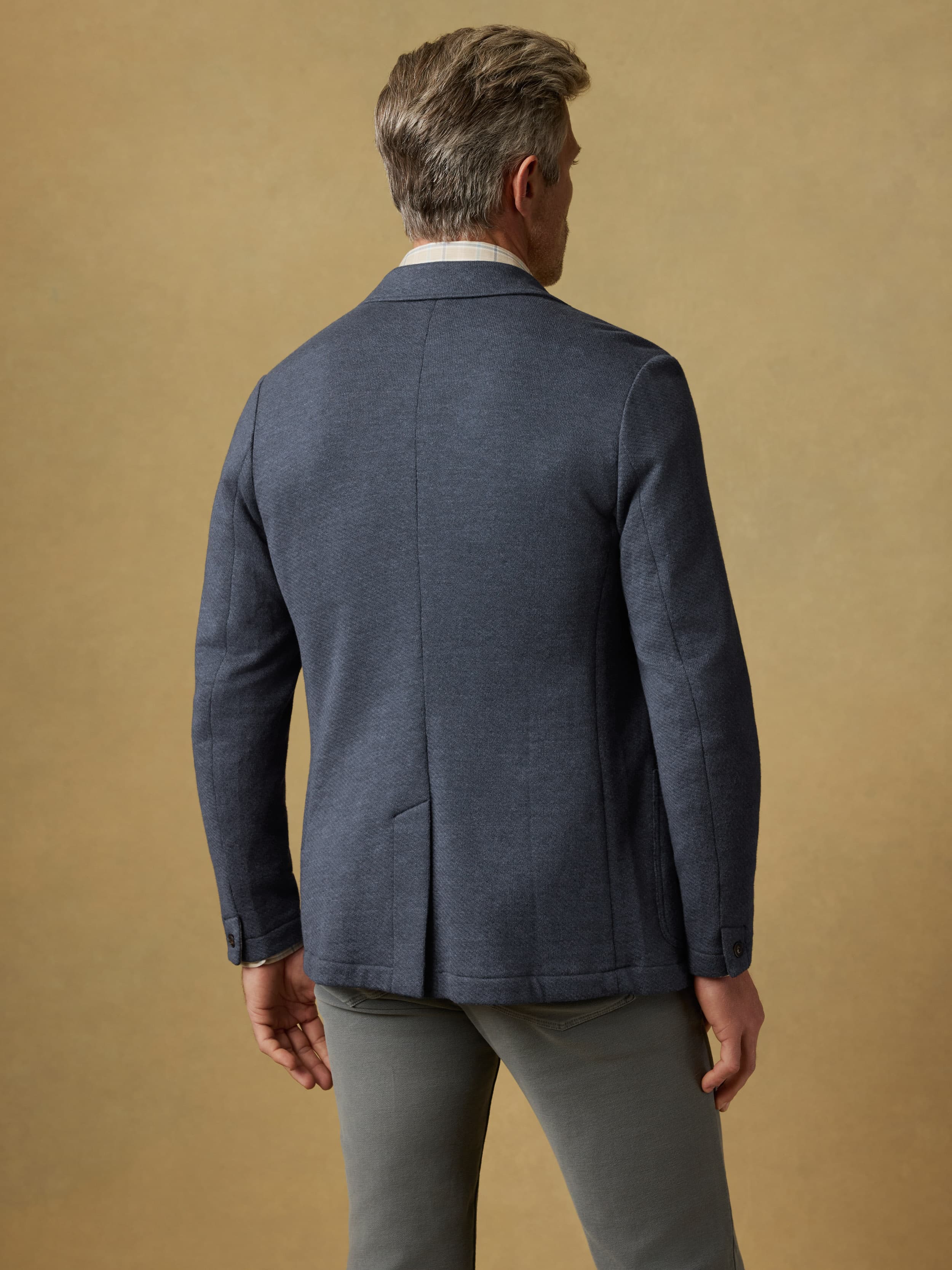 Back view of a man wearing a blue blazer and gray pants against a beige background