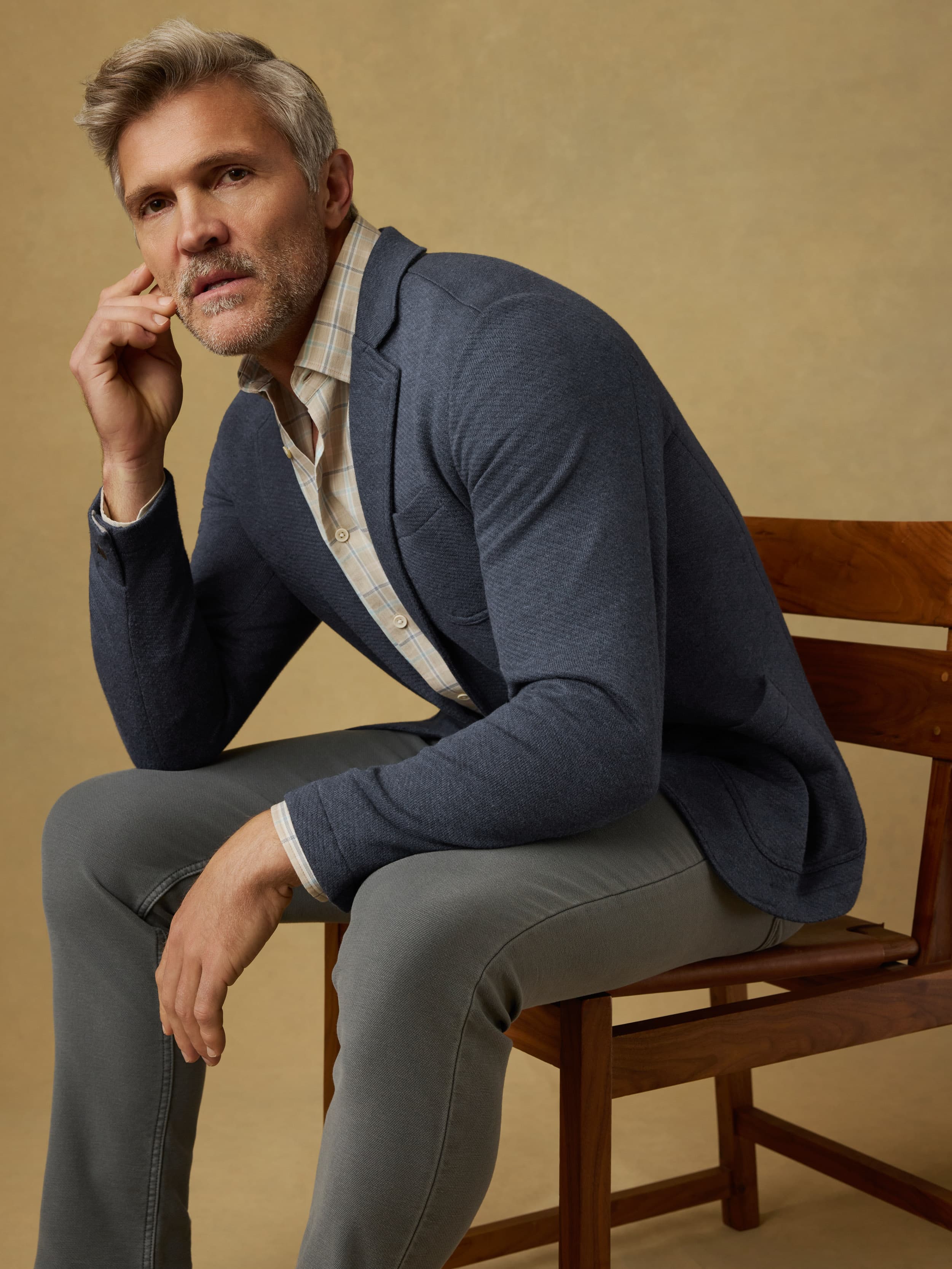 Man sitting on a wooden chair wearing a blue blazer over a plaid shirt with gray pants against a beige background