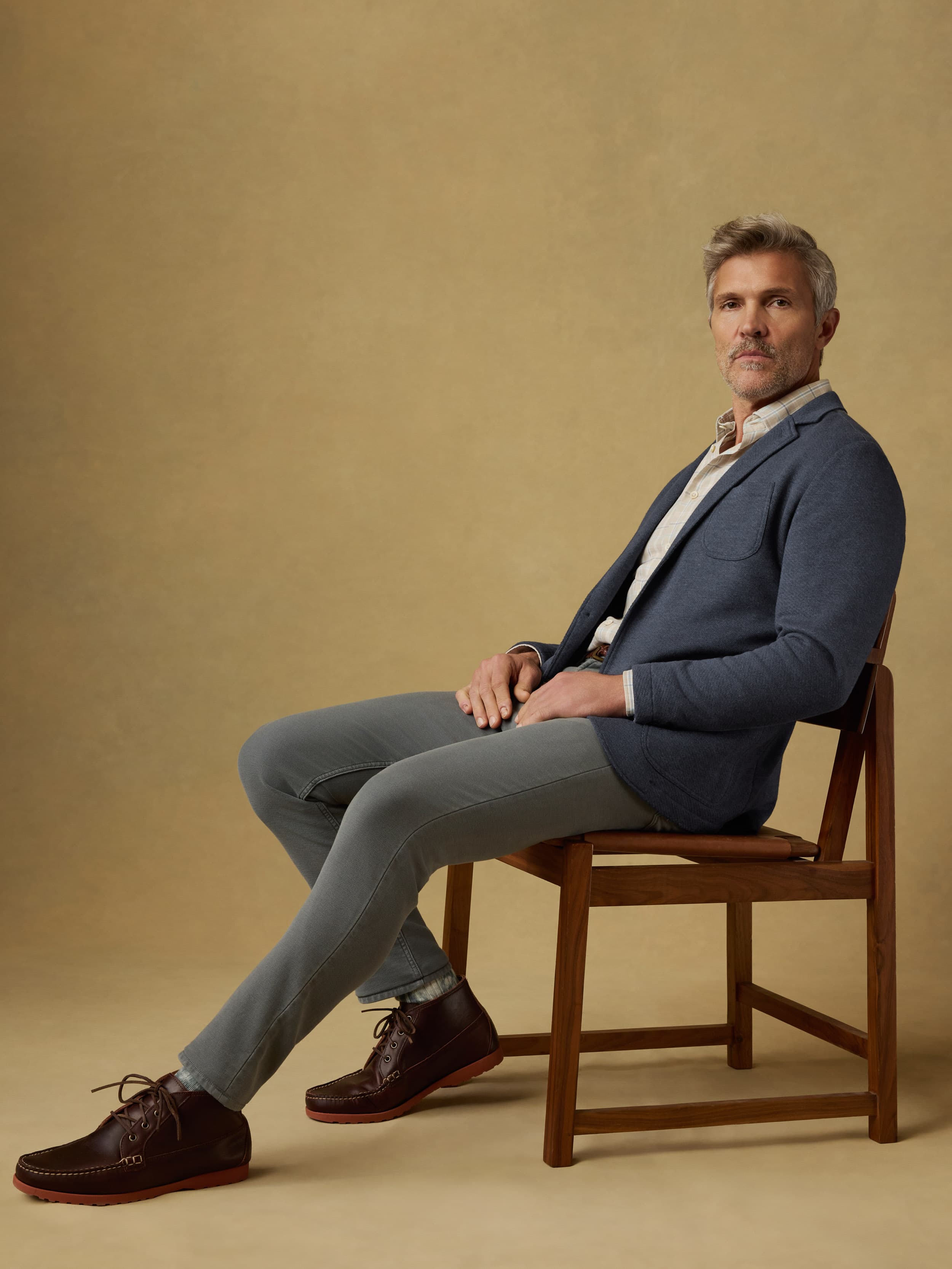 Man sitting on a wood chair wearing a blue blazer over a plaid shirt with gray pants against a beige background