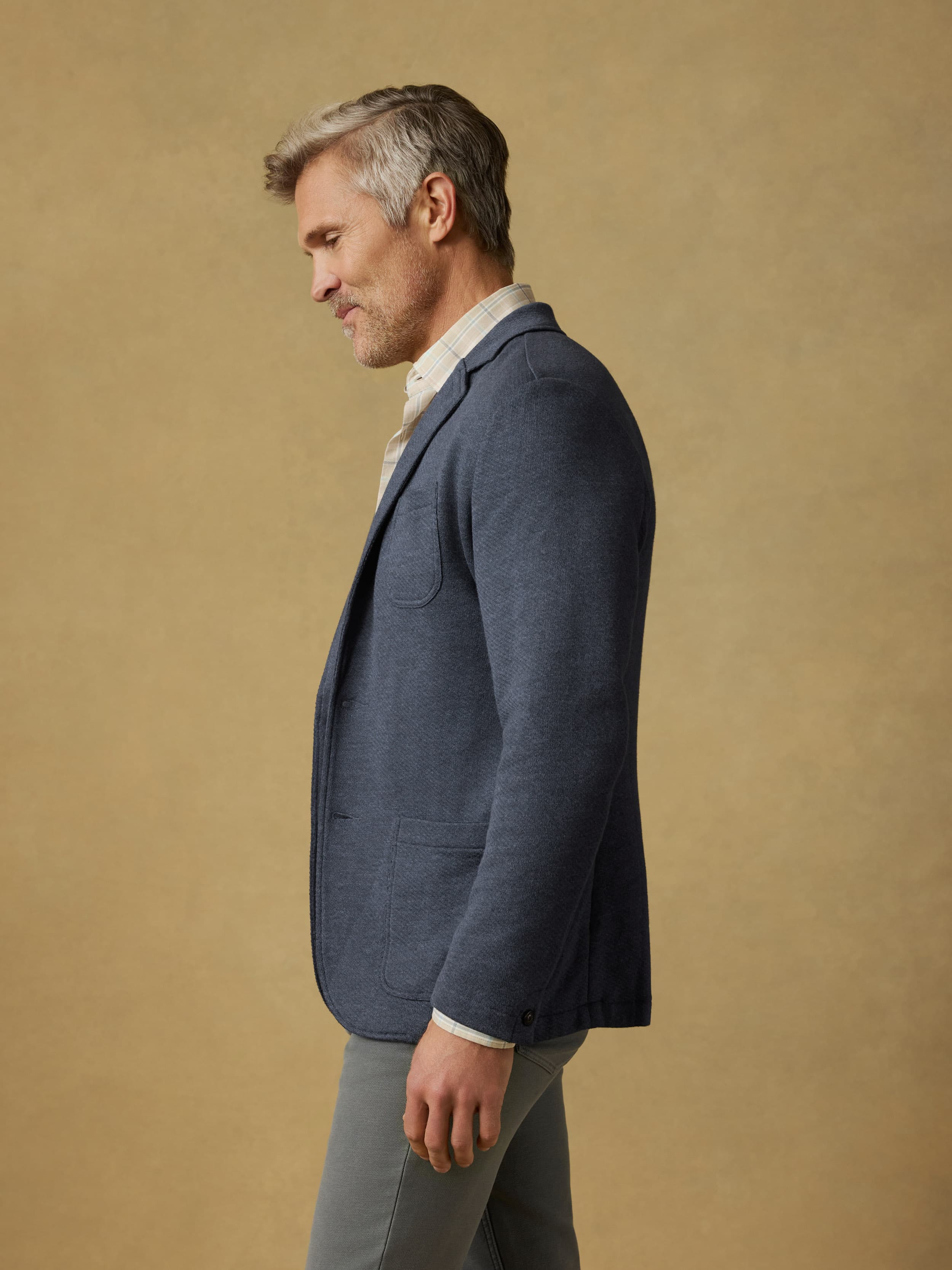 Man wearing a blue blazer over a plaid shirt with gray pants against a beige background