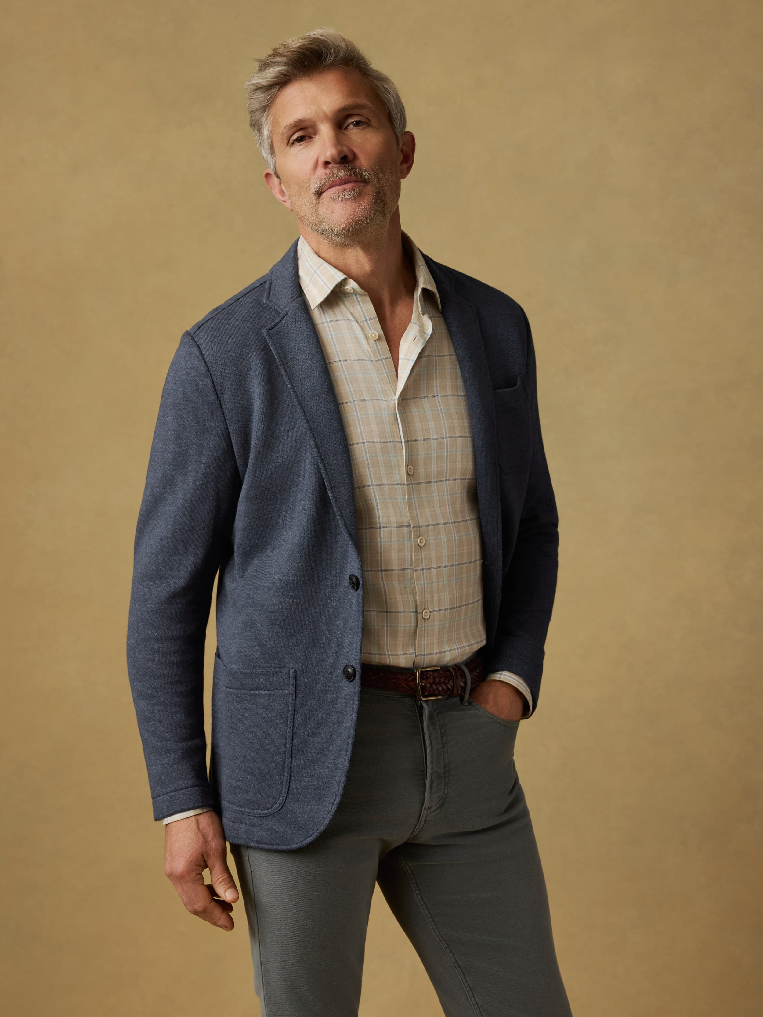 Man wearing a blue blazer over a plaid shirt with gray pants against a beige background