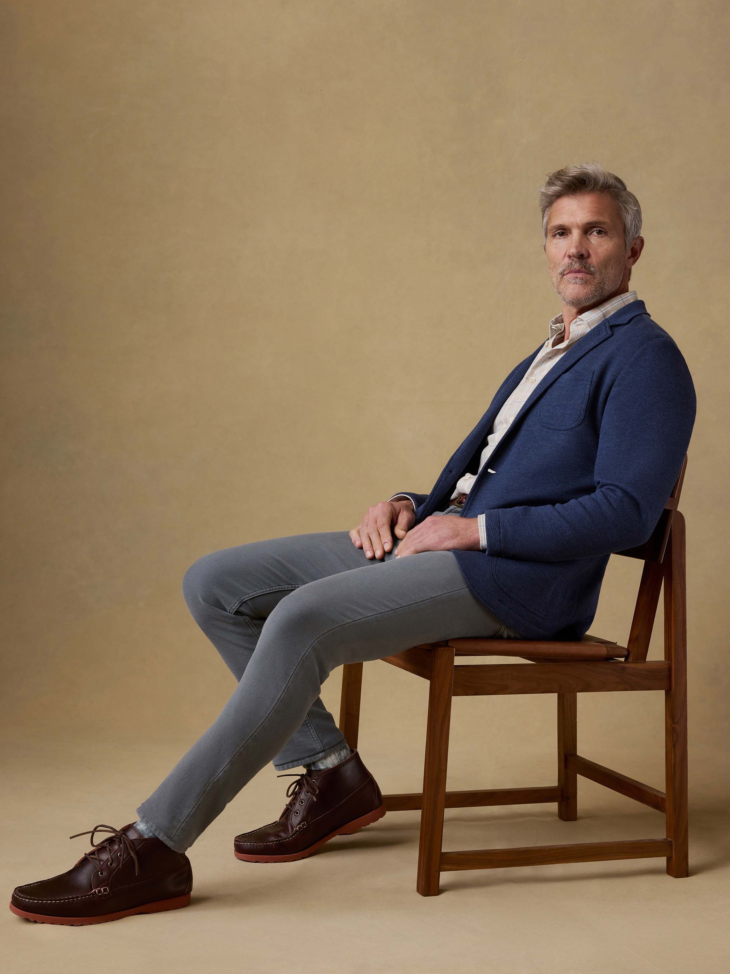 Man sitting on a wooden chair wearing a blue blazer and gray pants against a beige background