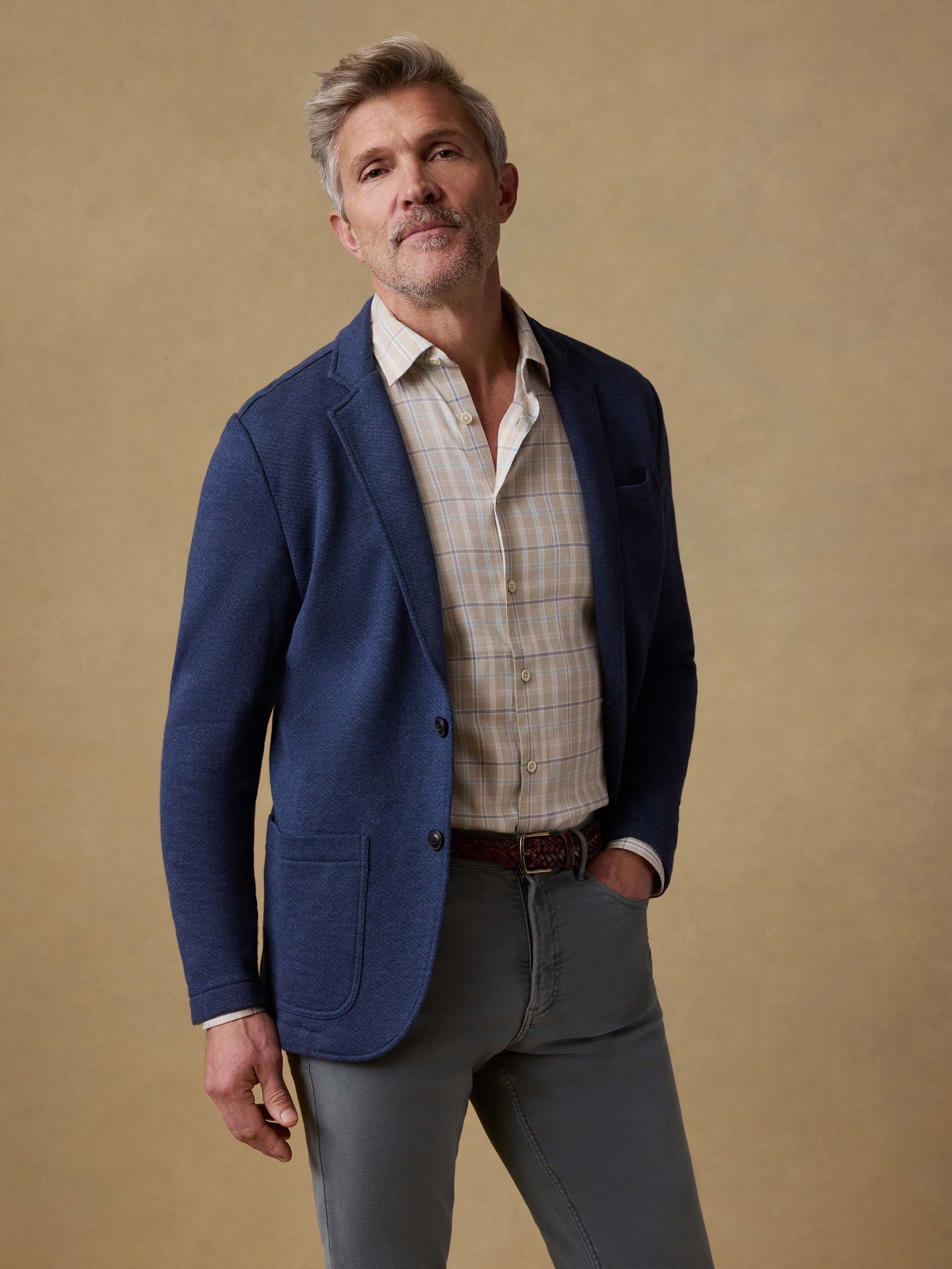 Man wearing a blue blazer over a plaid shirt with gray pants on a beige background