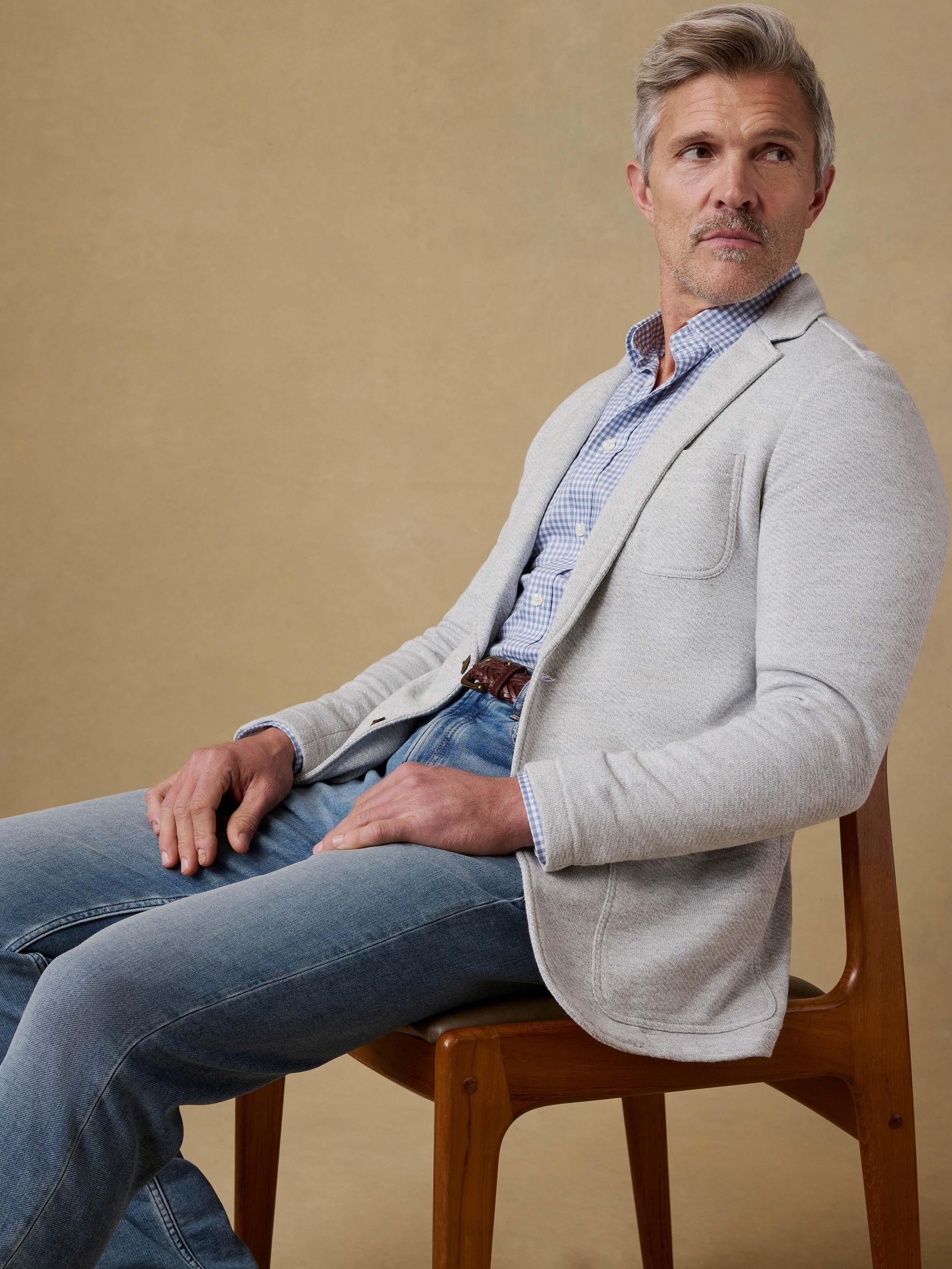 Man sitting on a wooden stool wearing a grey blazer over a blue checkered shirt and jeans against a beige background.