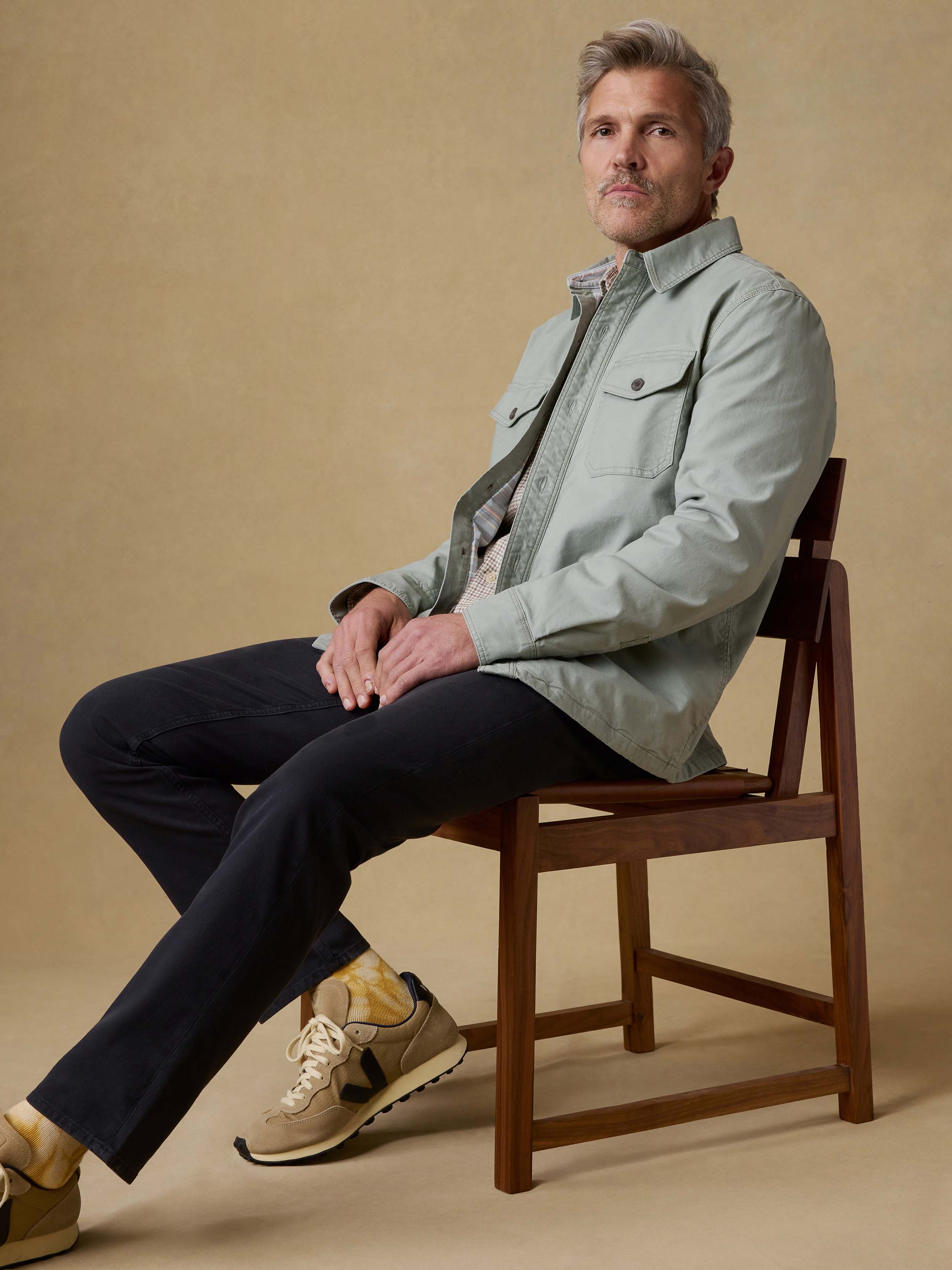 Man sitting on a wooden chair wearing a light green jacket and dark pants against a beige background
