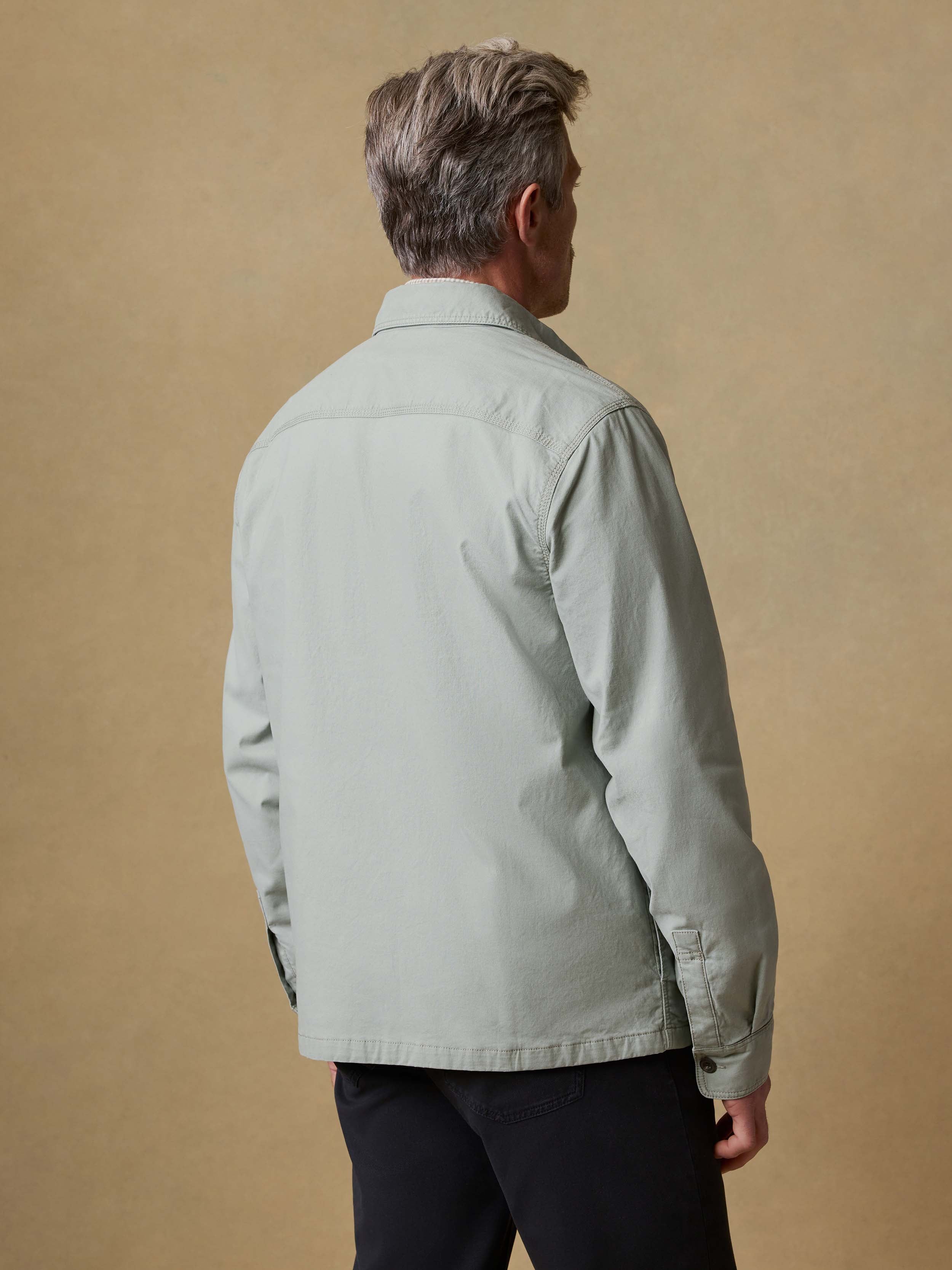 Back view of a man wearing a light green jacket and black pants against a beige background
