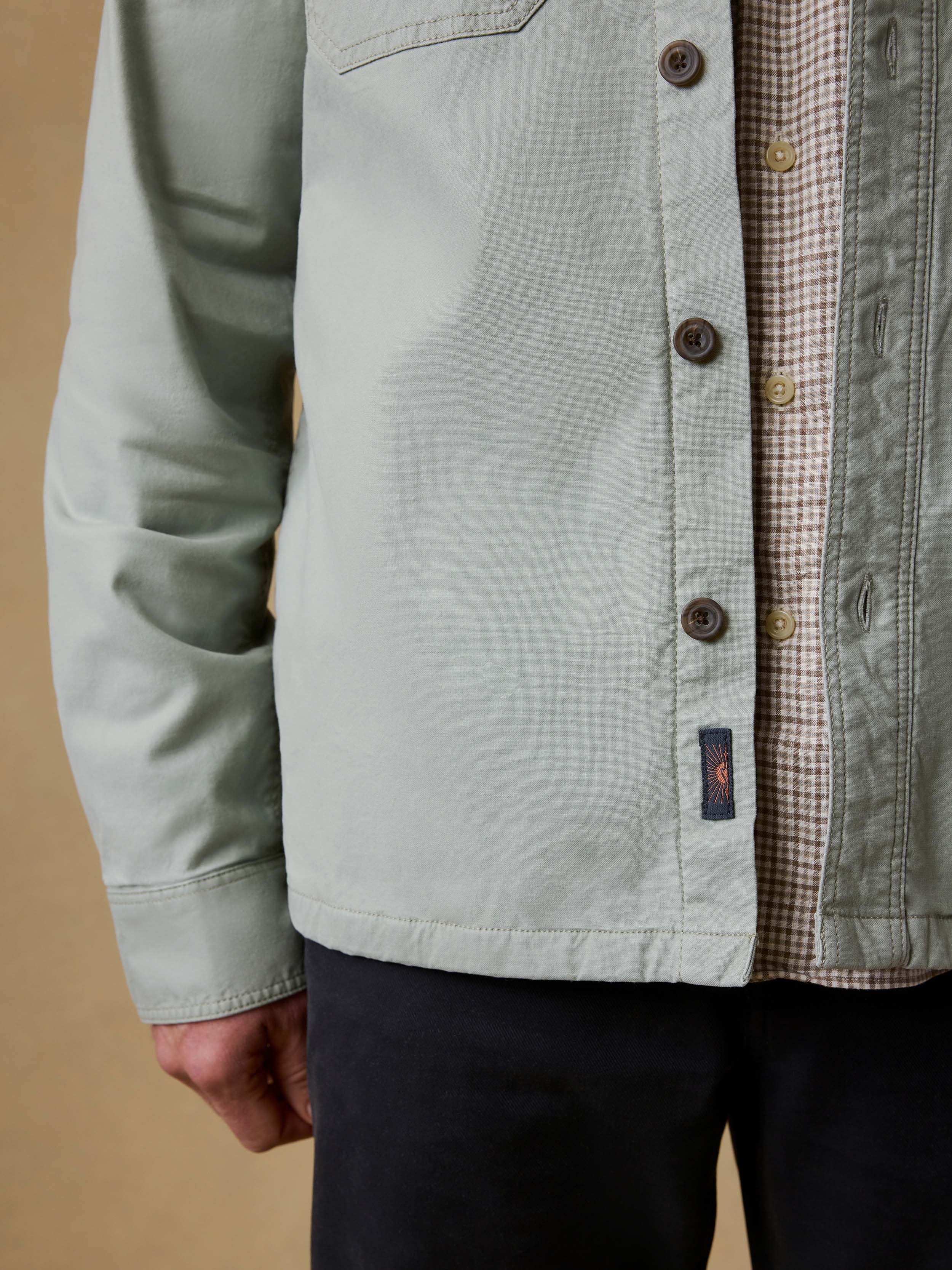 Detail shot of a man wearing a light green jacket over a plaid shirt and black pants against a beige background
