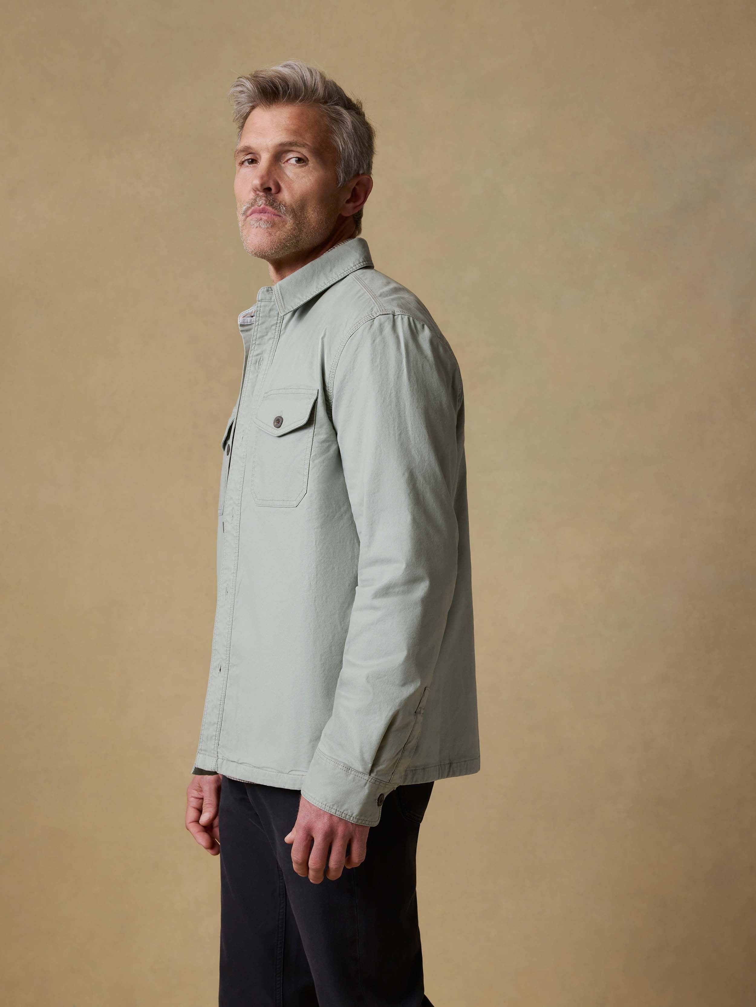 Man wearing a light green jacket over a plaid shirt and black pants against a beige background