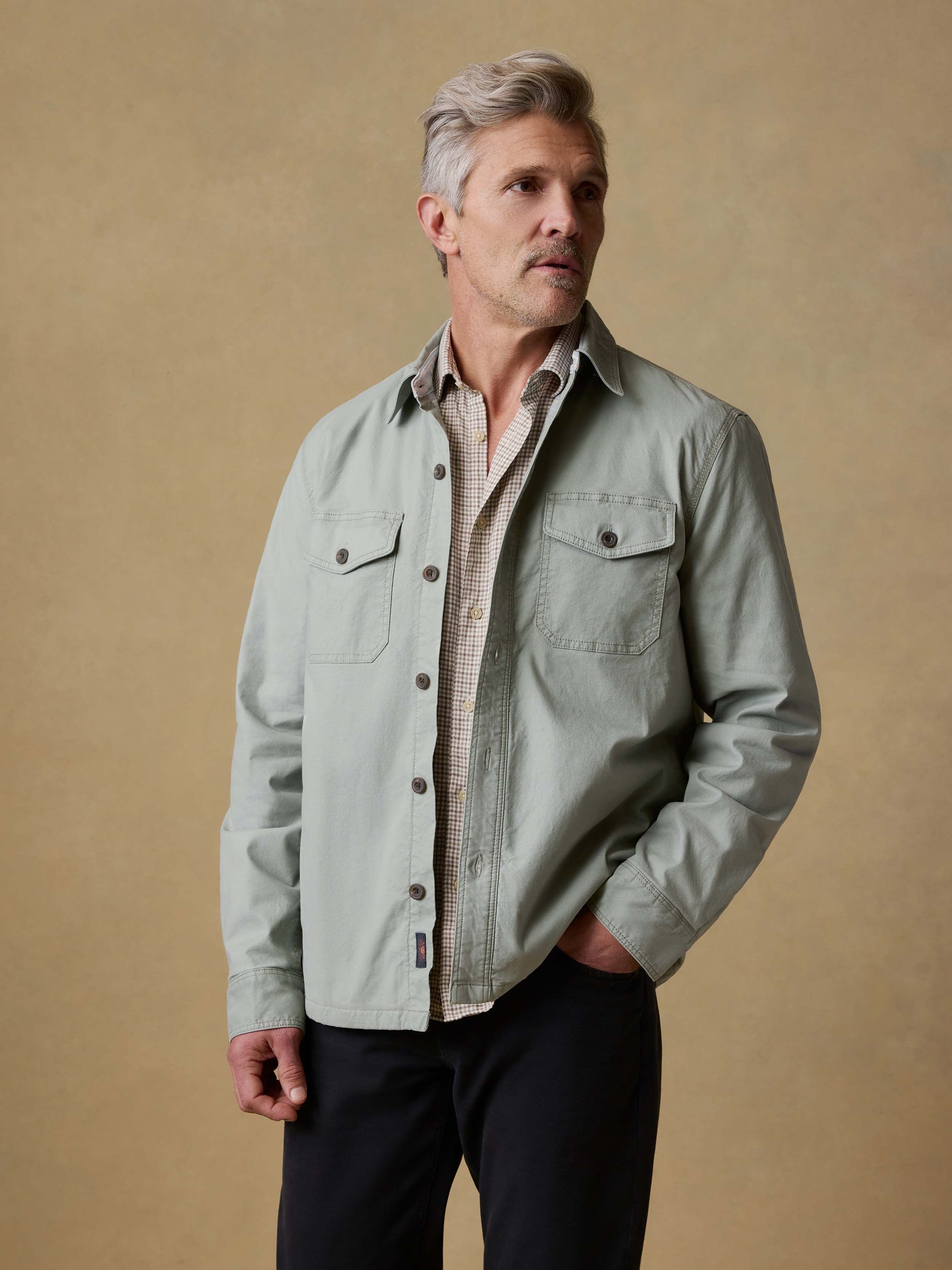 Man wearing a light green jacket over a plaid shirt and black pants against a beige background