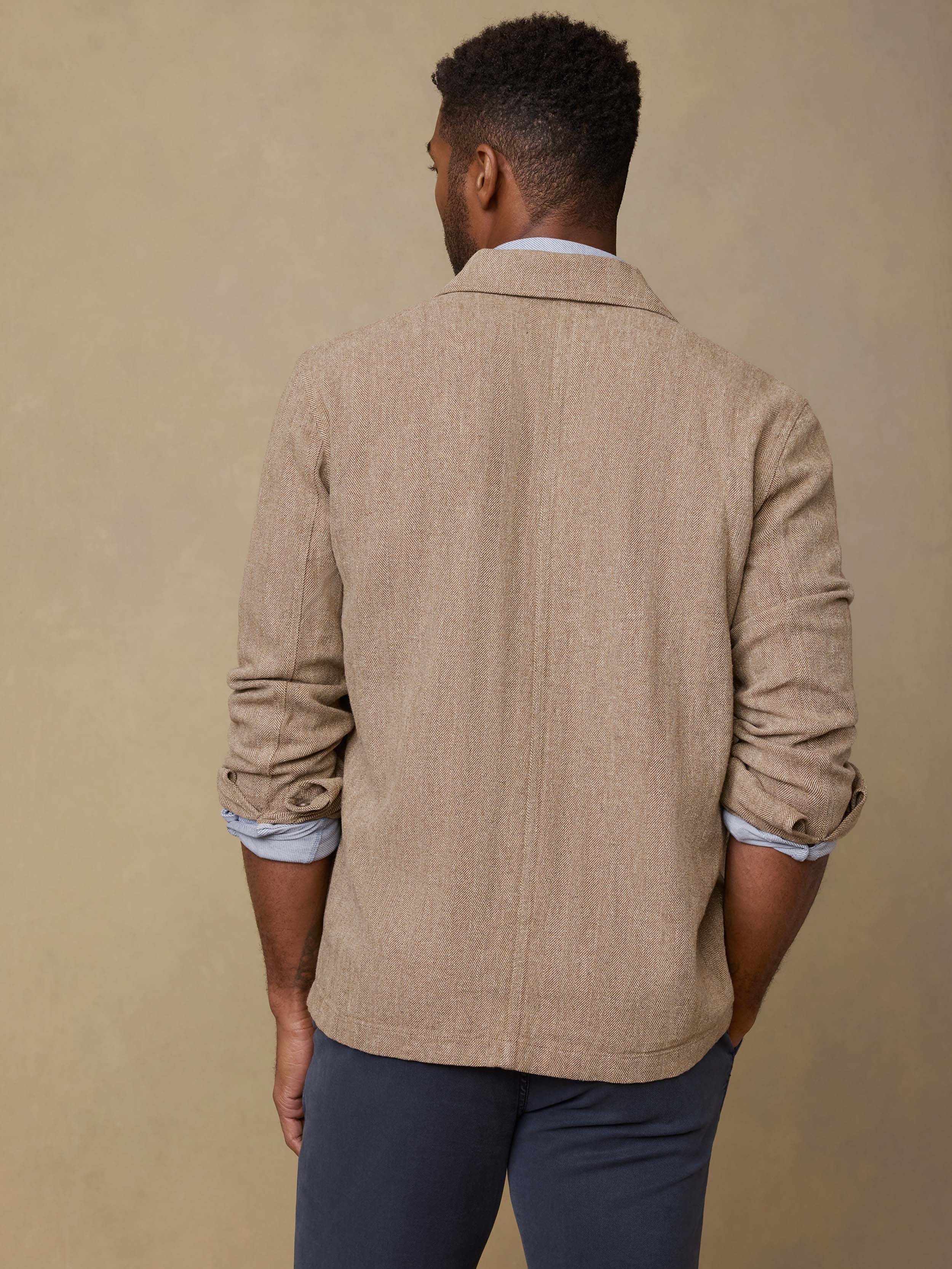 Back view of a man wearing a beige blazer over a light blue shirt with rolled-up sleeves on a beige background