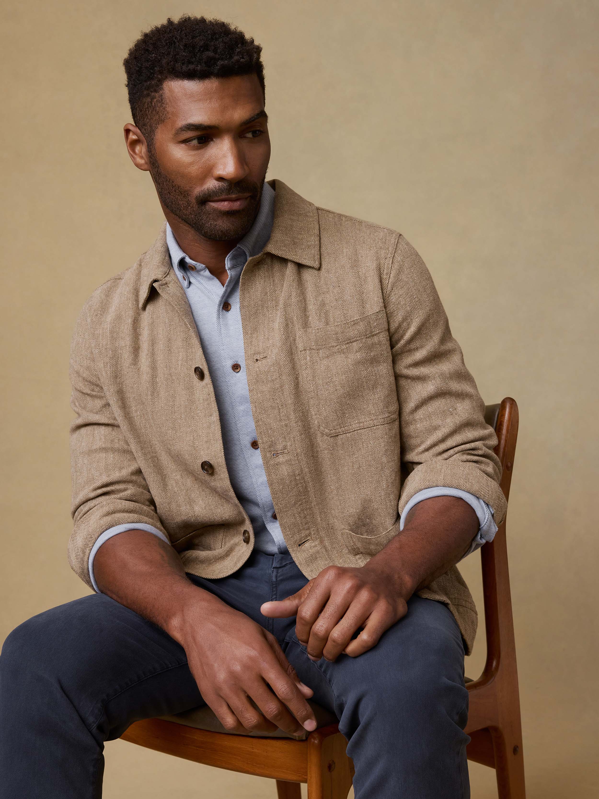 Man sitting on a wooden chair wearing a beige jacket over a light blue shirt.