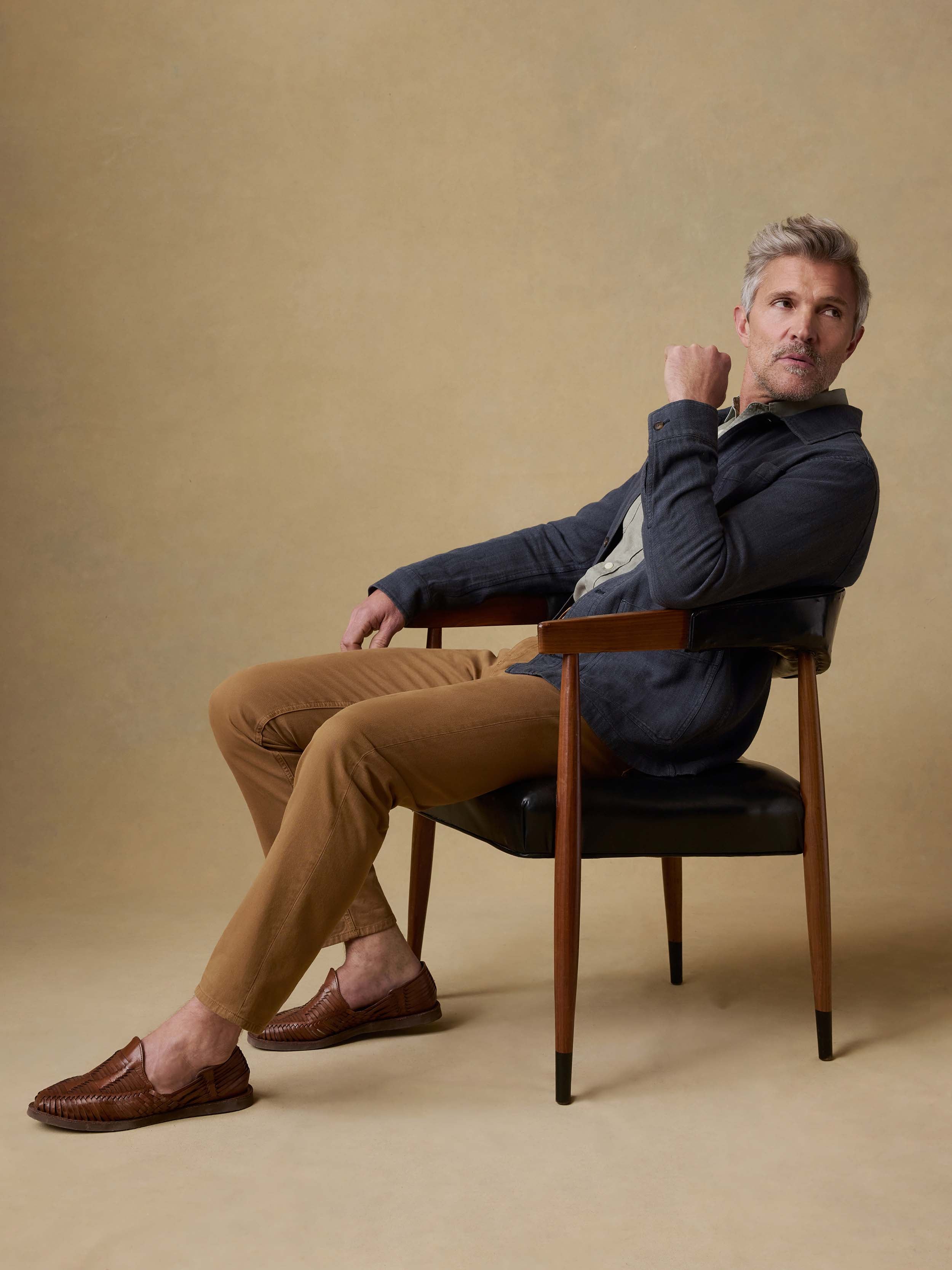 Man sitting on a chair wearing a blue sweater and brown pants against a beige background