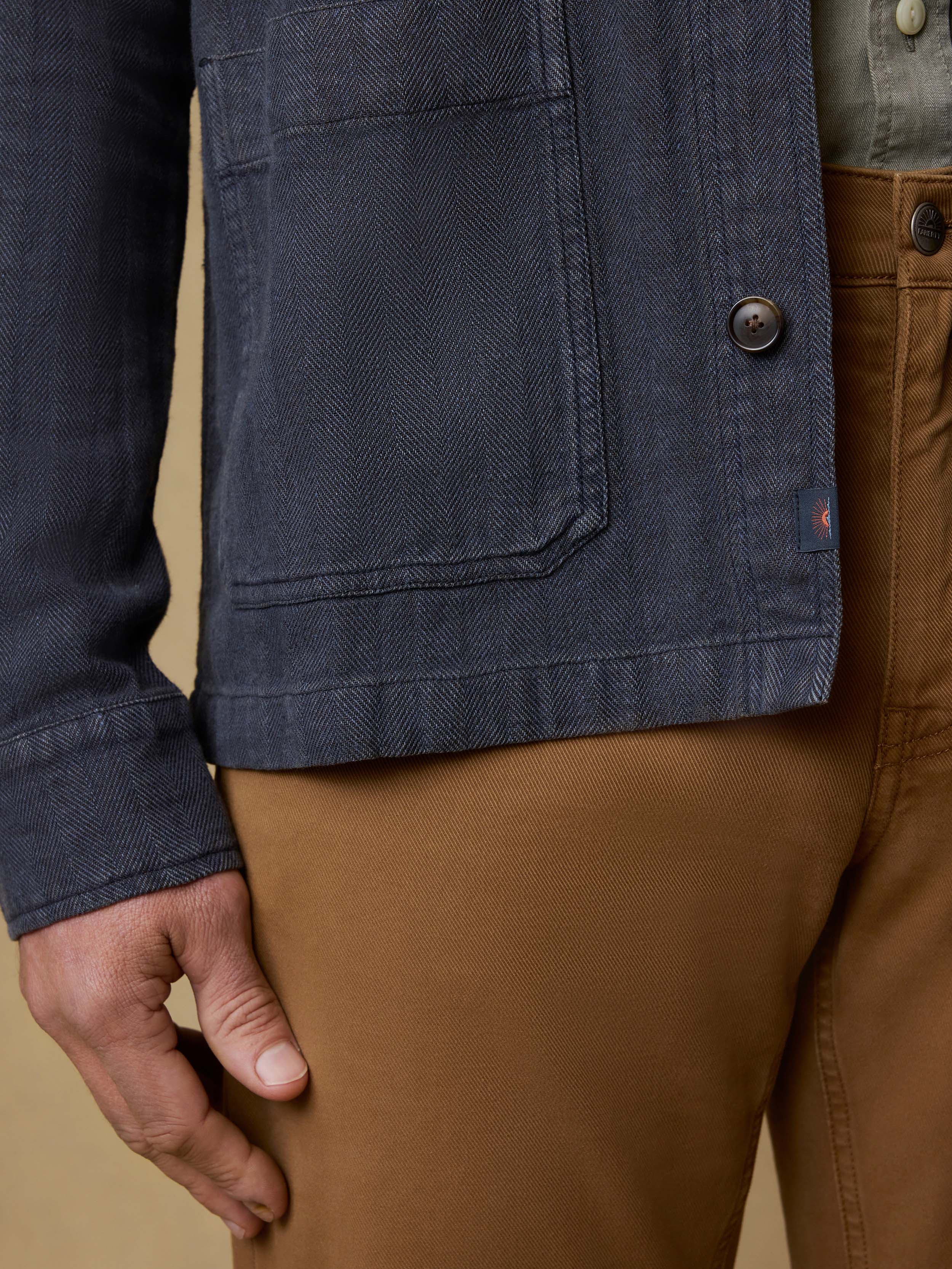 Close-up of a person wearing a dark blue jacket with a button and brown pants on a beige background.