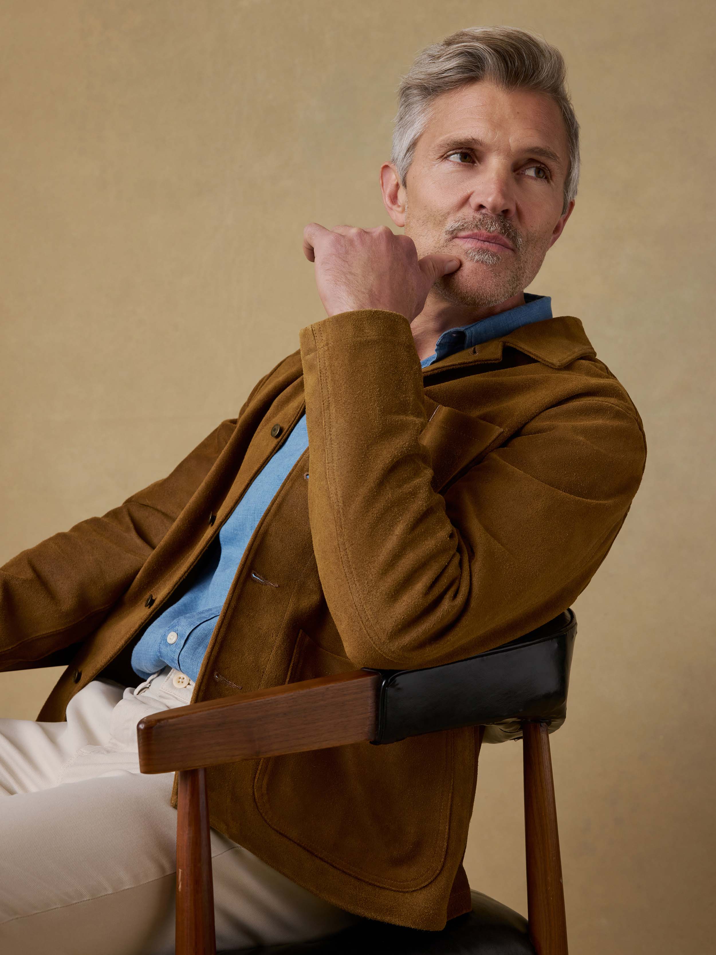 Man sitting on a chair wearing a brown suede jacket and blue shirt against a beige background