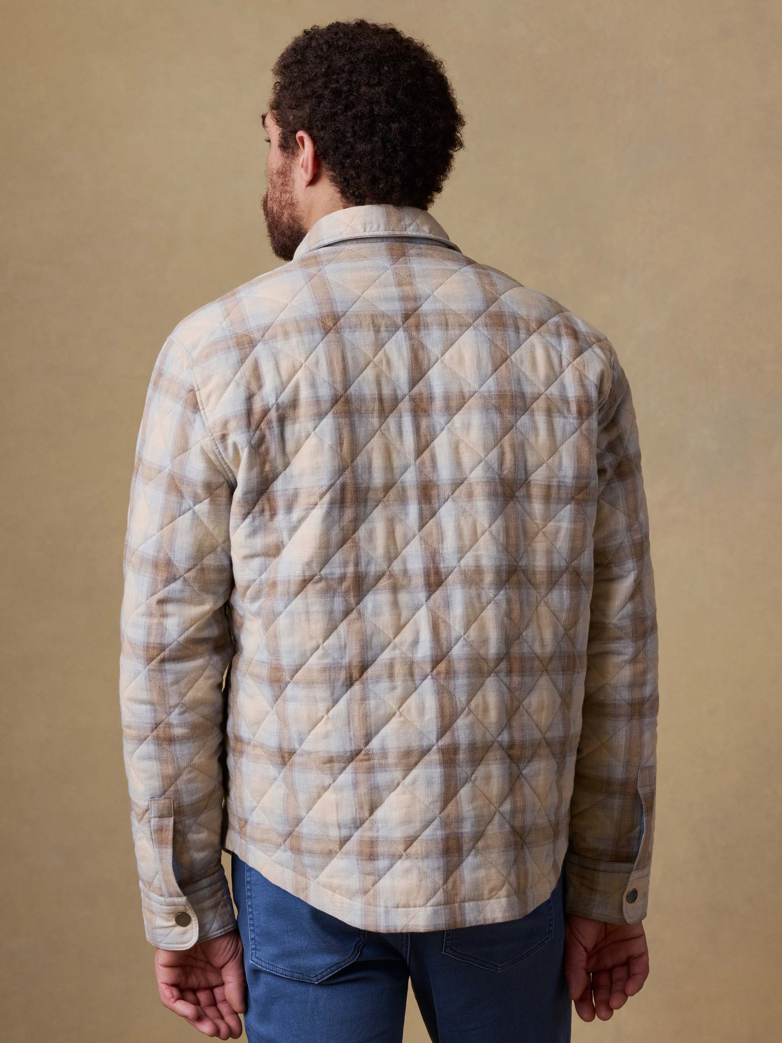 Back view of a man wearing a plaid shirt jacket and blue jeans against a beige background