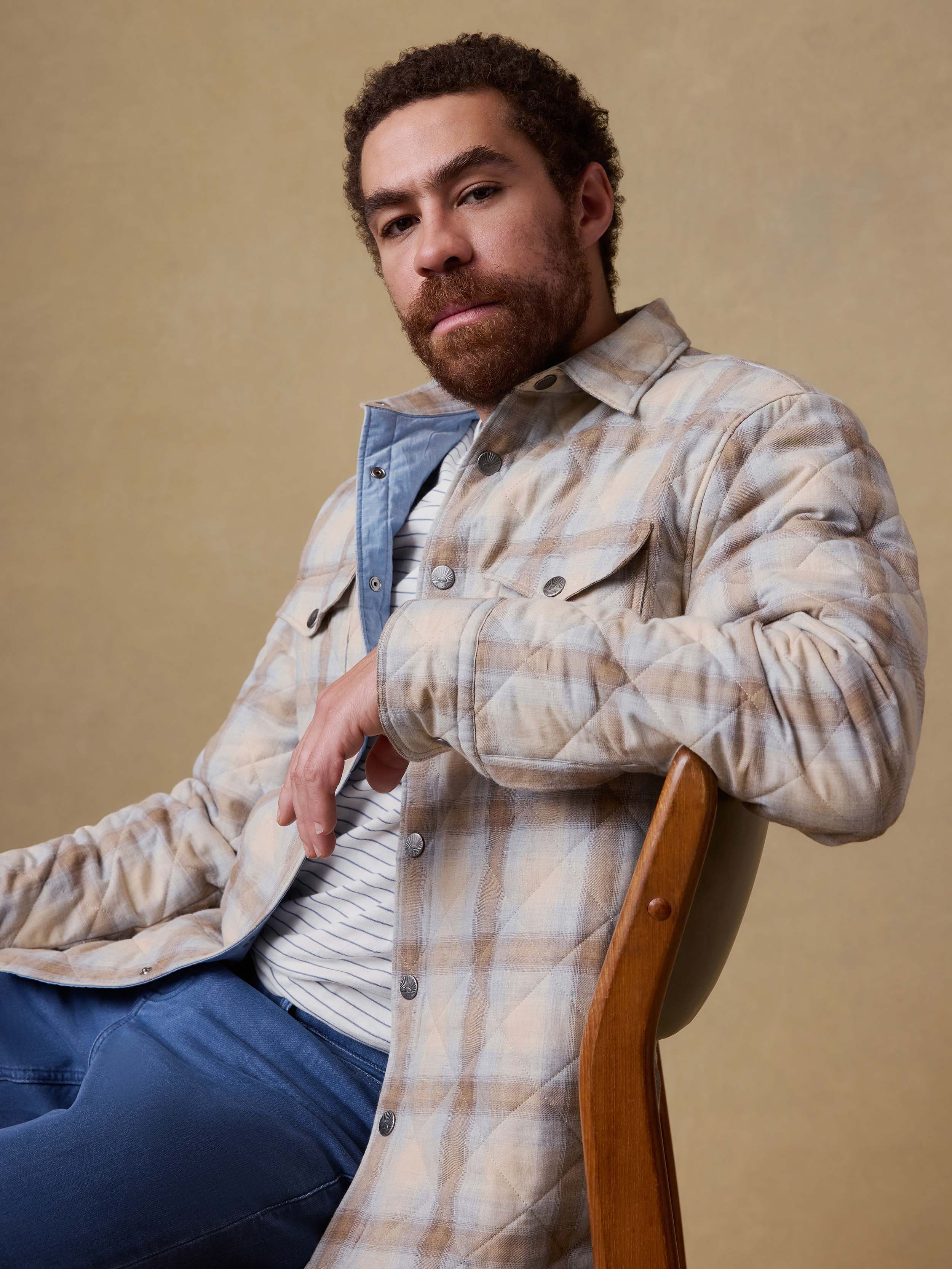 Man sitting on a chair wearing a plaid shirt jacket over a striped shirt and blue jeans against a beige background