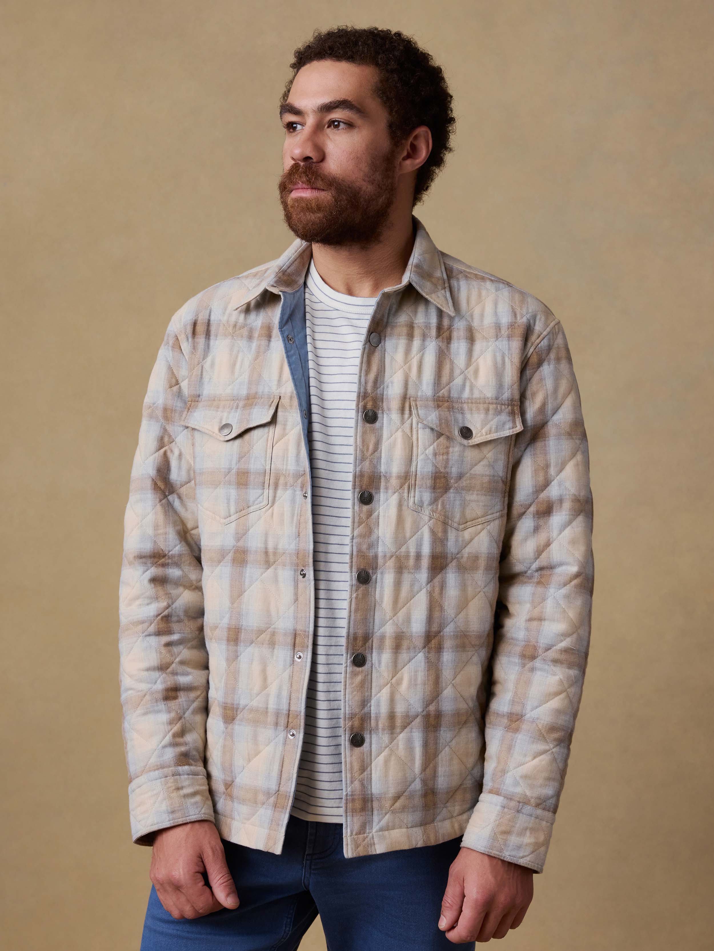Man wearing a plaid shirt jacket over a striped shirt and blue jeans against a beige background