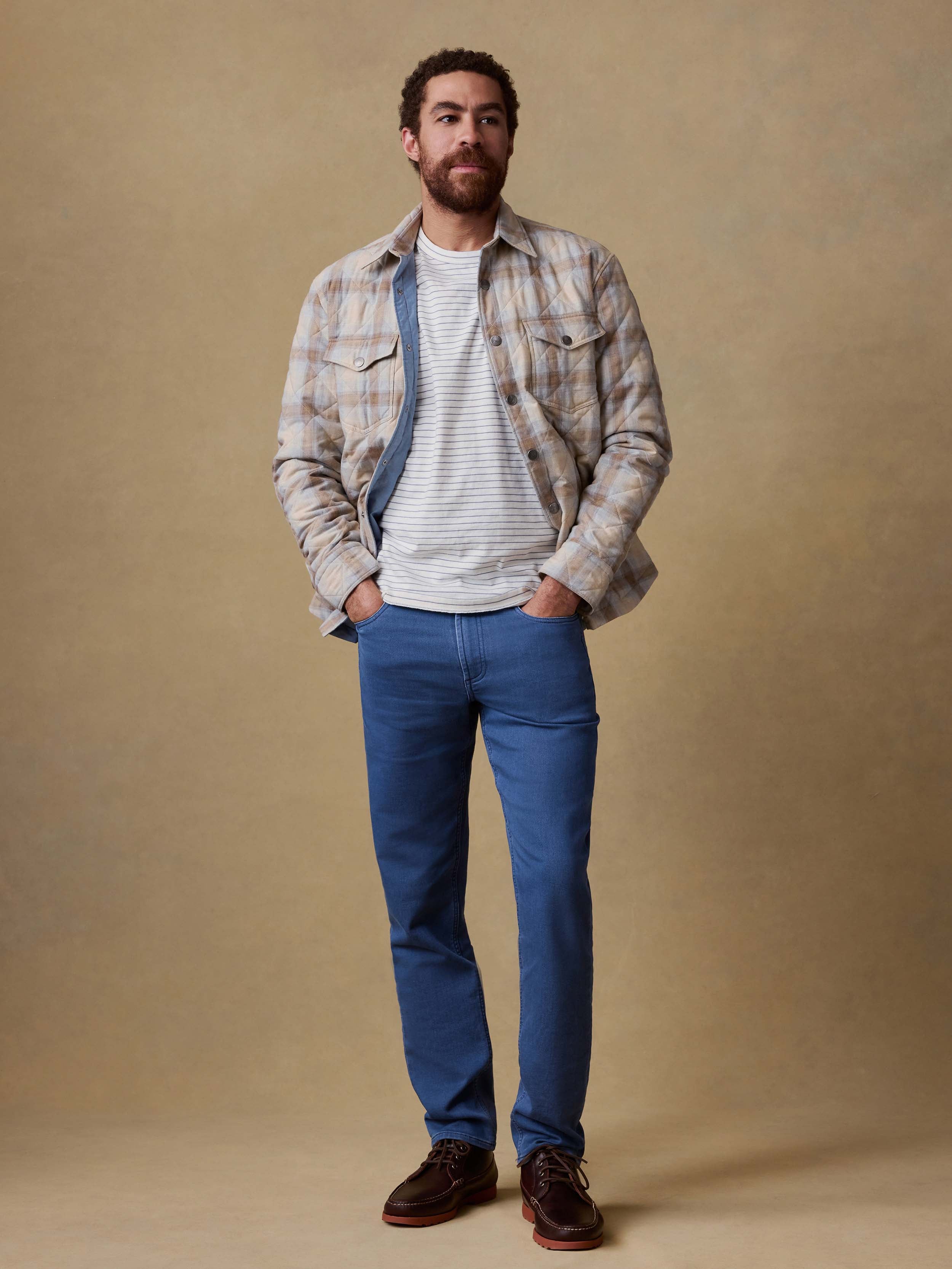 Man wearing a plaid shirt jacket over a striped shirt and blue jeans against a beige background