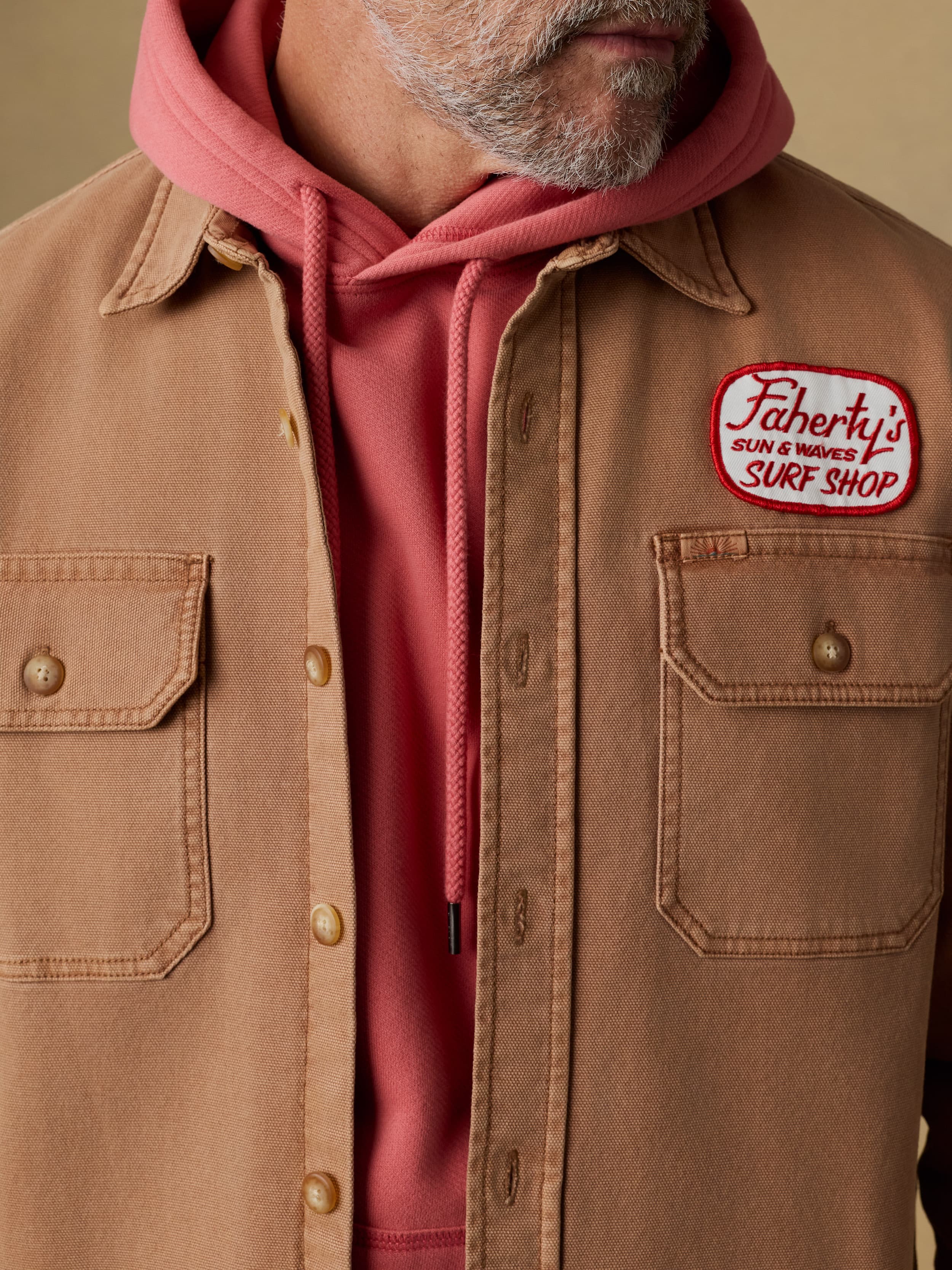 Man wearing a brown jacket with a Faherty patch at the left chest over a red hoodie against a beige background