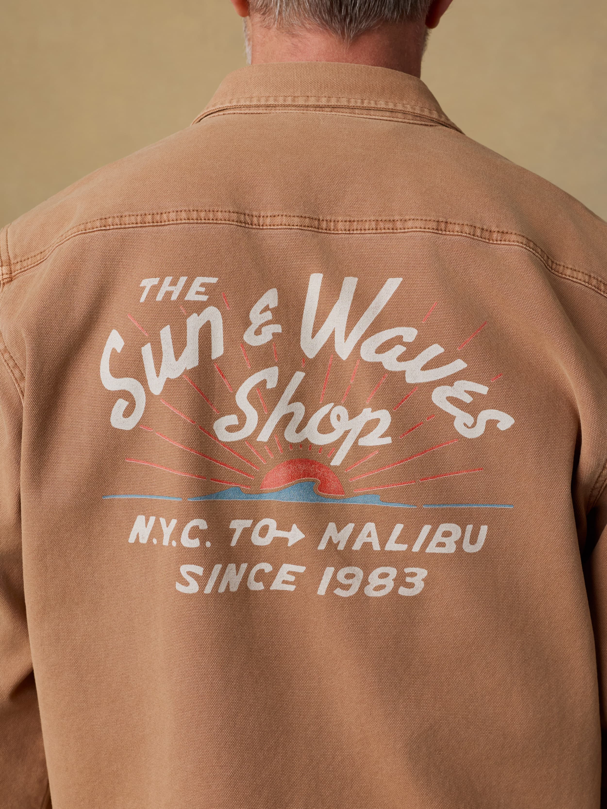 Back view of a man wearing a brown jacket with 'The Sun & Waves Shop' Faherty graphic on the back against a beige background