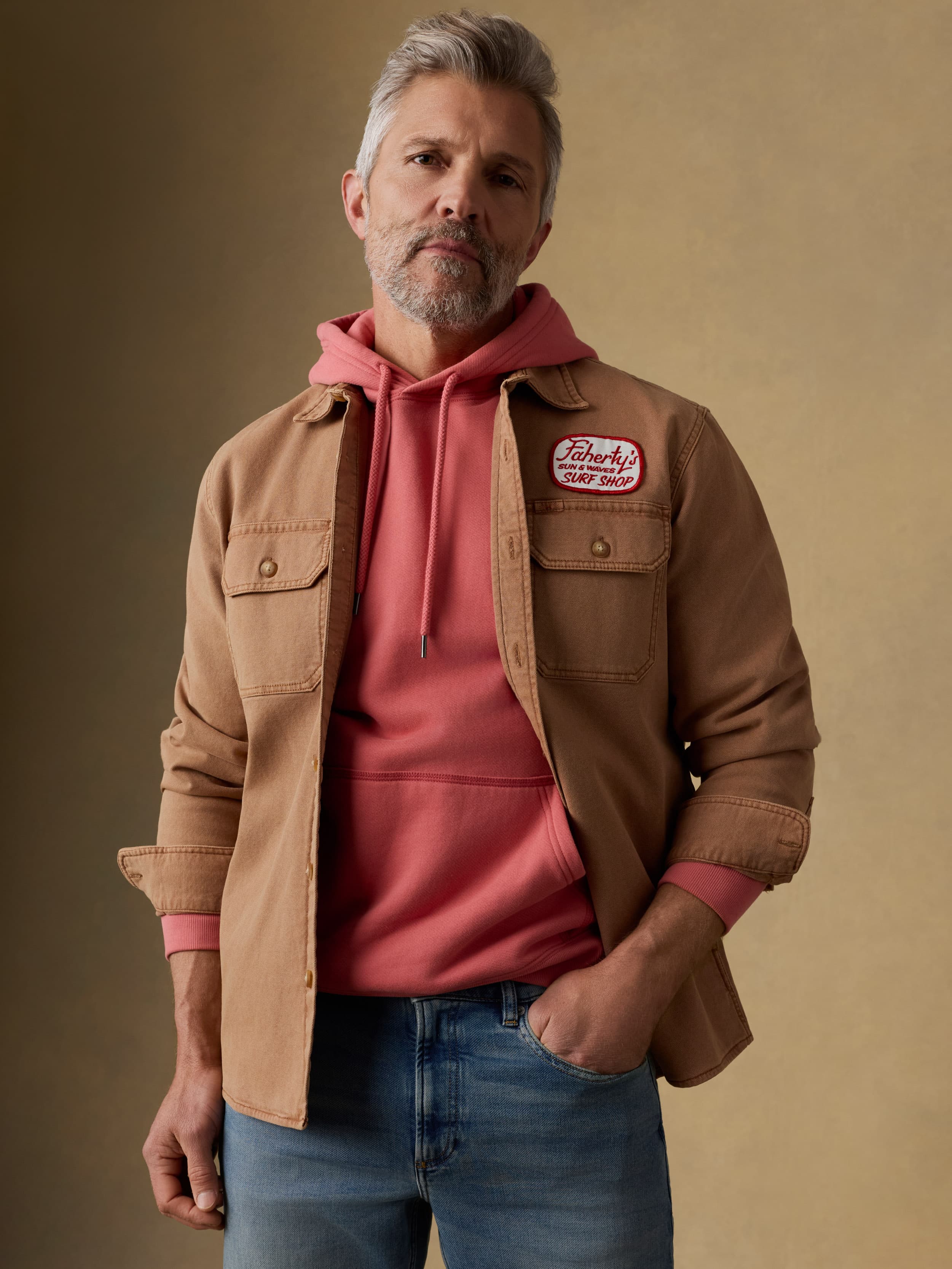 Man wearing a brown jacket with a Faherty patch at the left chest over a red hoodie against a beige background