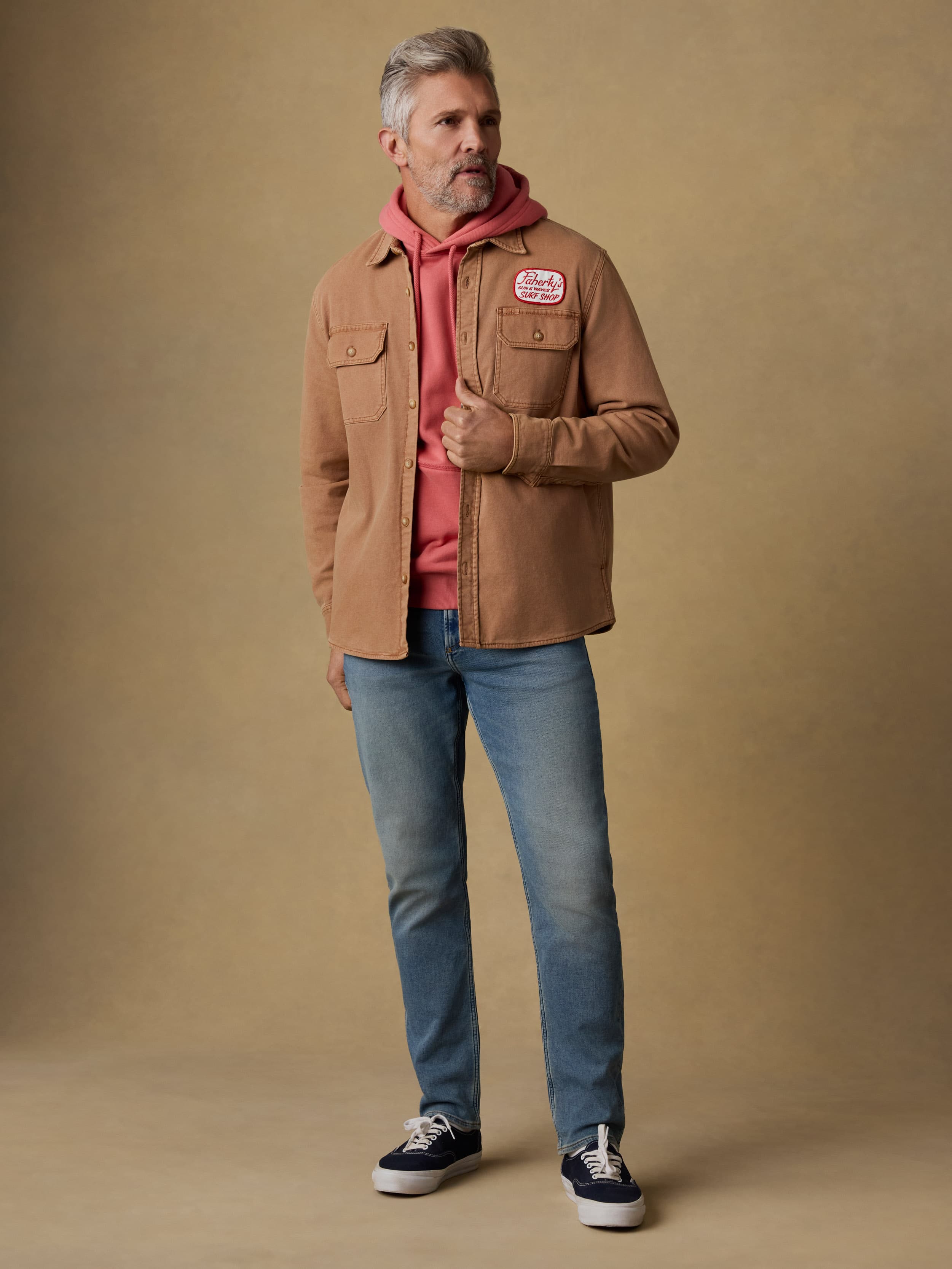 Man wearing a brown jacket with a Faherty patch at the left chest over a red hoodie against a beige background