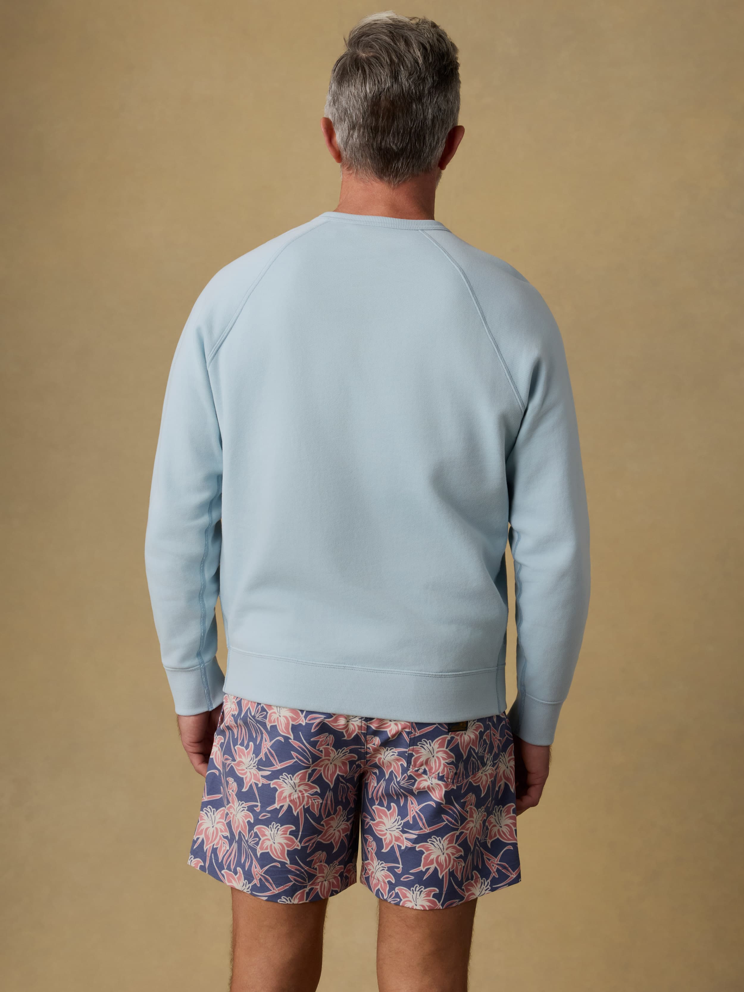 Back view of a man wearing a light blue crewneck sweatshirt and floral shorts against a beige background