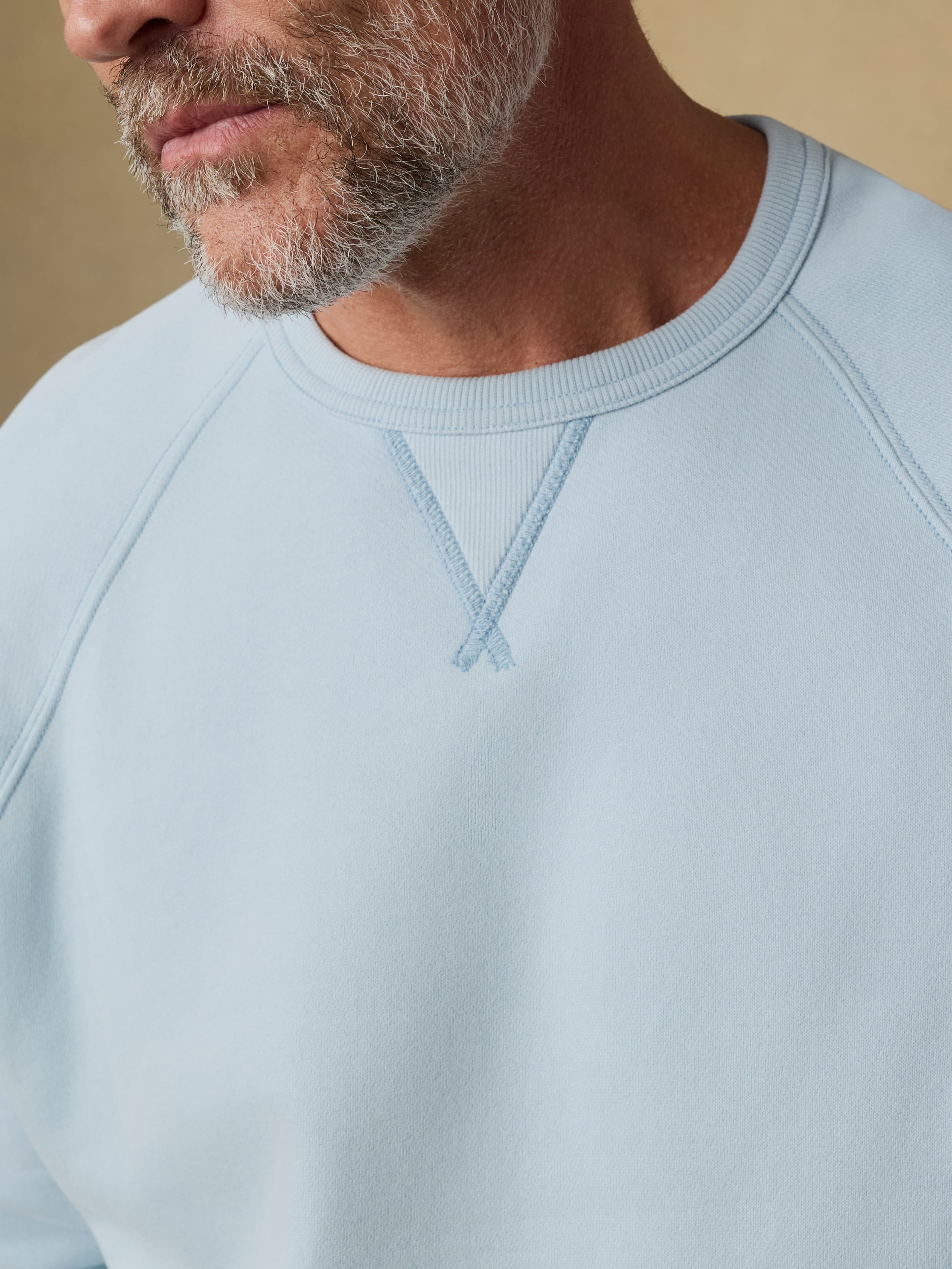 Close up shot of a man wearing a light blue crewneck sweatshirt against a beige background