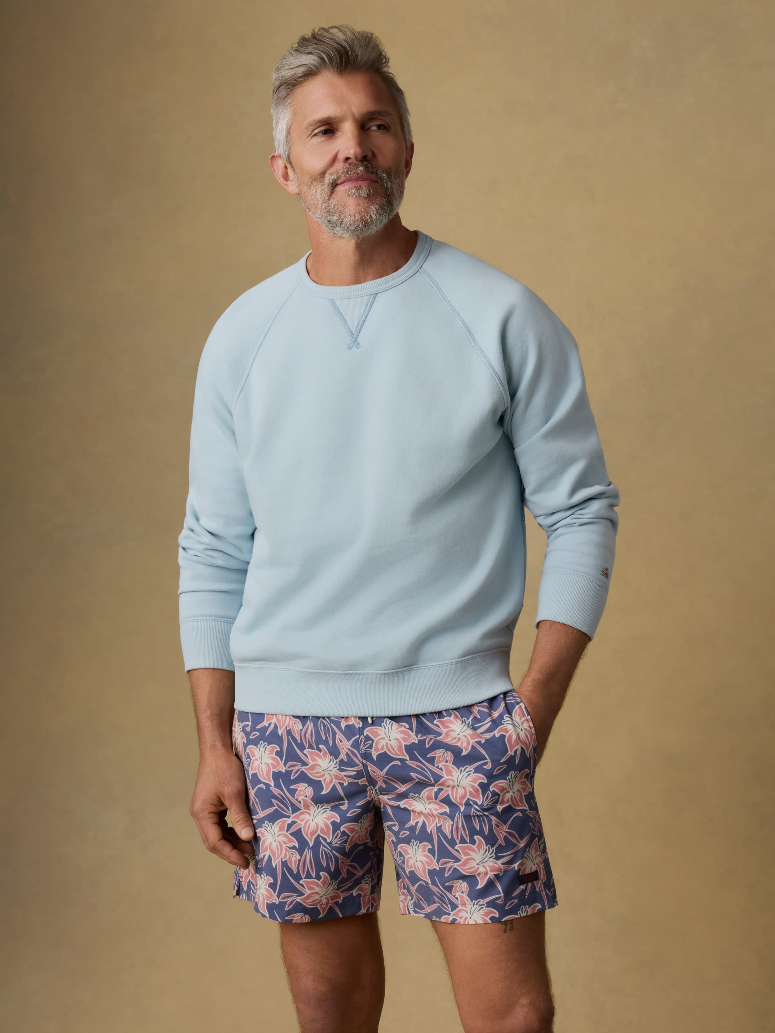 Man wearing a light blue crewneck sweatshirt and floral shorts against a beige background