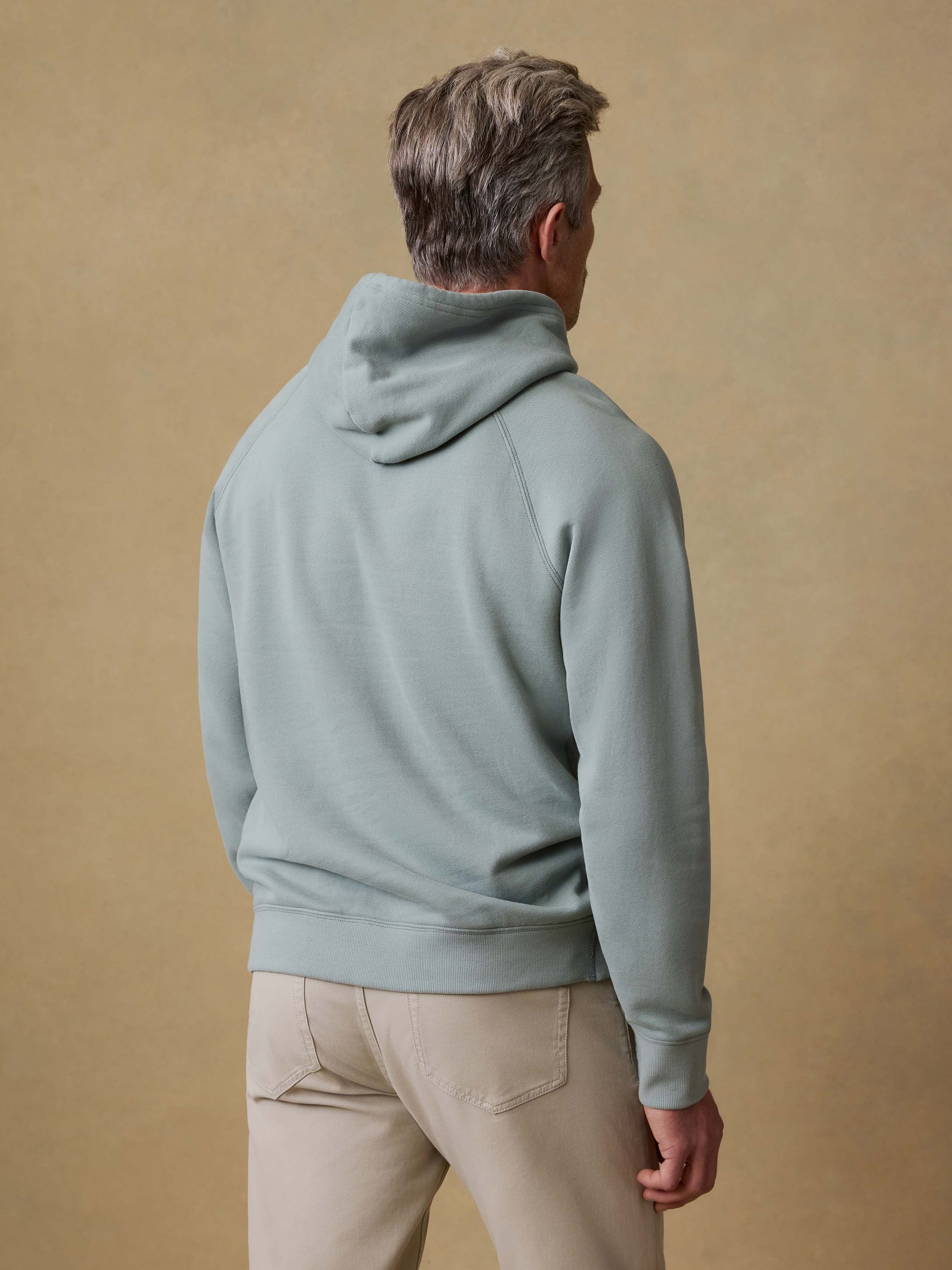 Back view of a man is wearing a light green hoodie and beige pants against a beige background