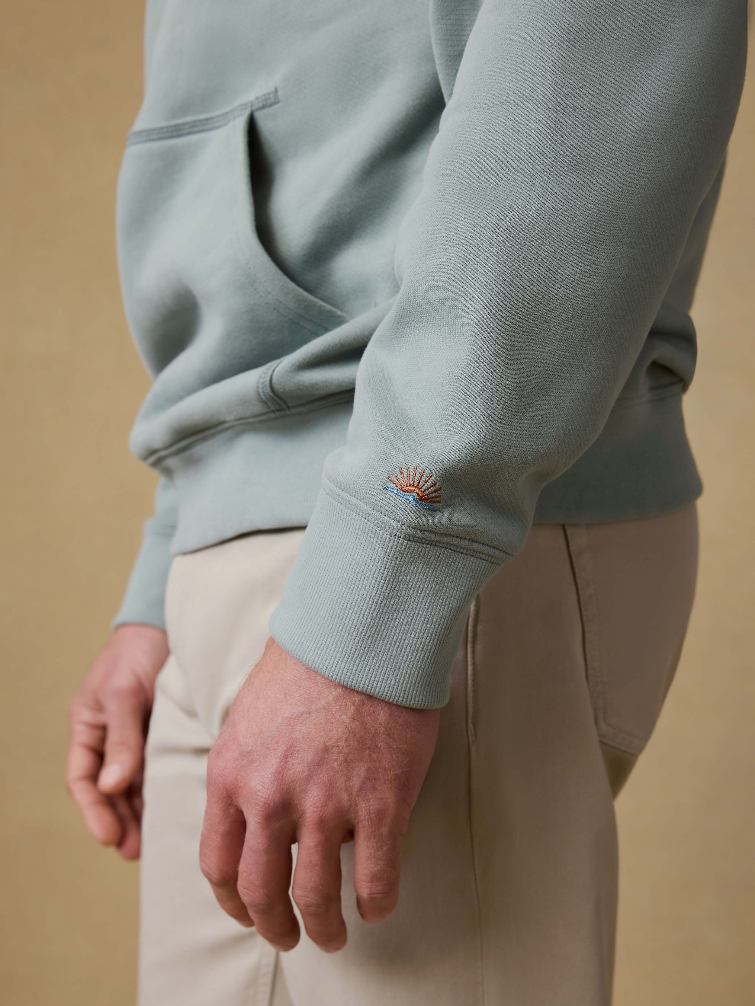 Man wearing a light green sweatshirt with an embroidered Faherty logo on the sleeve against a beige background